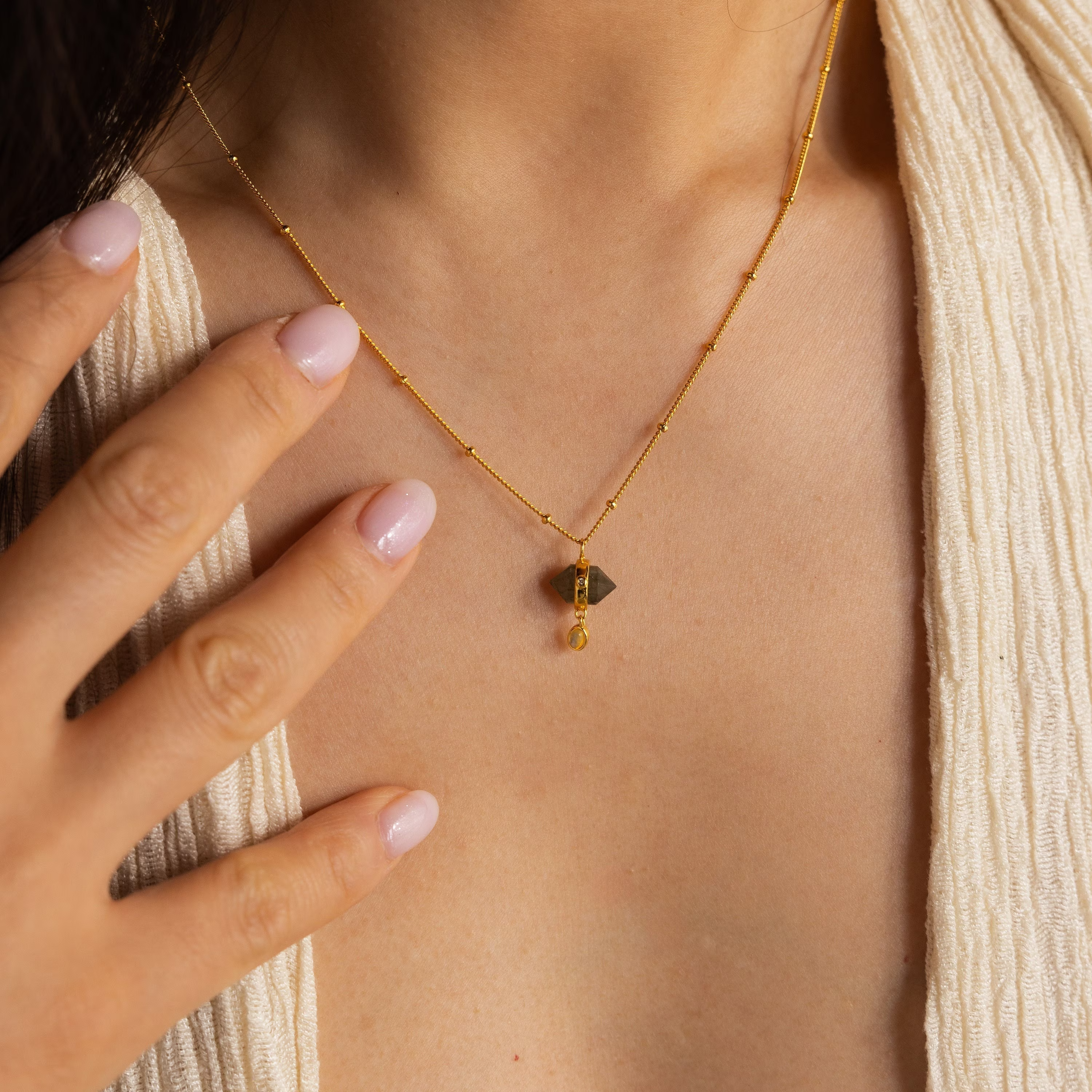 A woman wears the Labradorite & Moonstone Drop Necklace, its brown stone pendant resting on her chest as she touches it with manicured nails.
