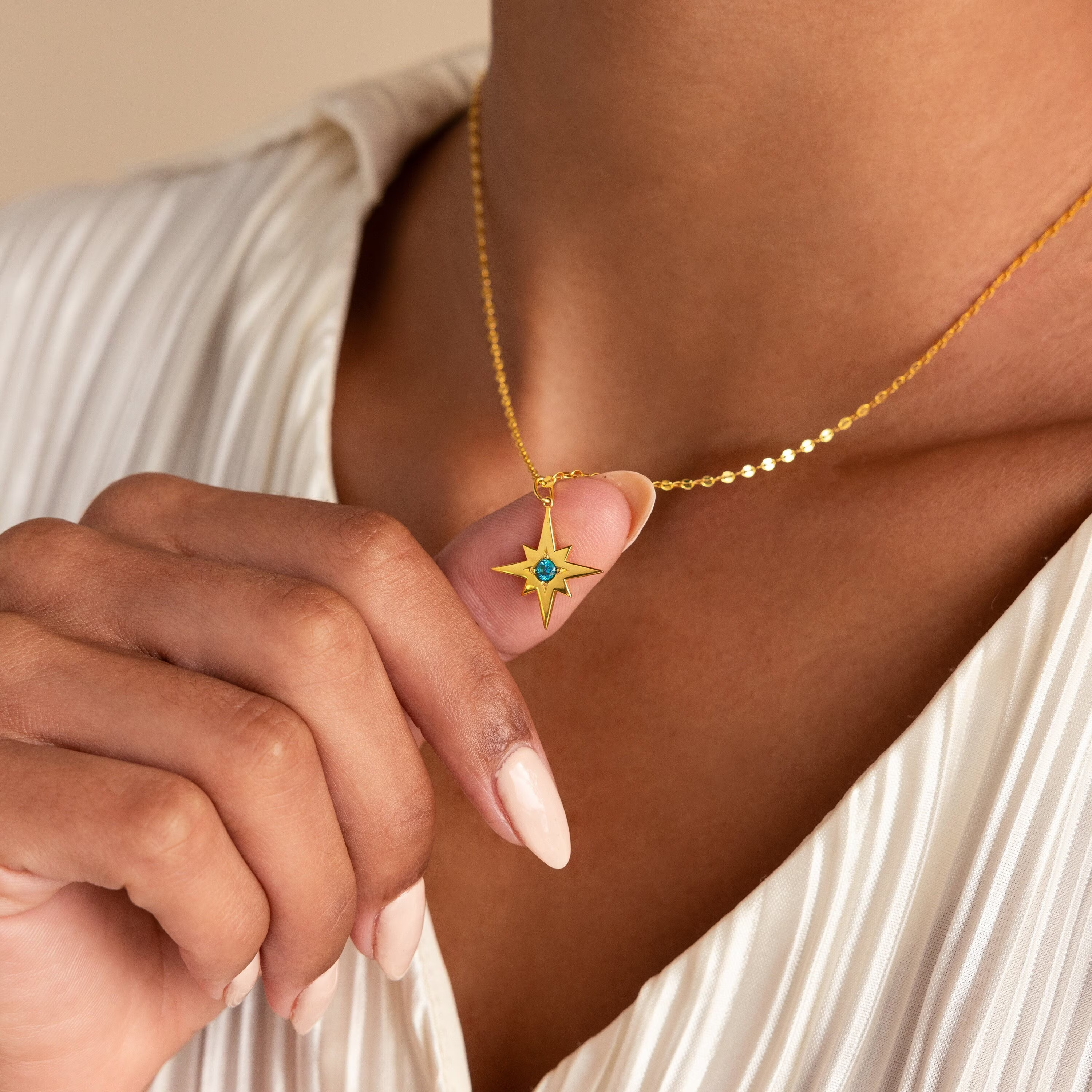 A person holds the Star Birthstone Necklace—a celestial gold star pendant with a blue gemstone—while wearing a cream blouse, making it an ideal personalized gift.