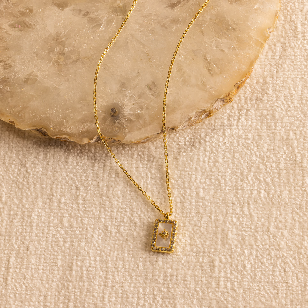 The Pave Pearl North Star Necklace, a gold piece with a rectangular pendant inspired by celestial jewelry, is shown on a textured surface with a stone background.