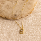 The Pave Pearl North Star Necklace, a gold piece with a rectangular pendant inspired by celestial jewelry, is shown on a textured surface with a stone background.