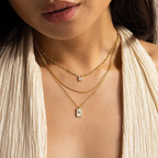 A woman wears the Pave Pearl North Star Necklace layered with gold necklaces and rectangular pendants over a cream textured top, shown from the shoulders up—an ideal style for those who love celestial jewelry.