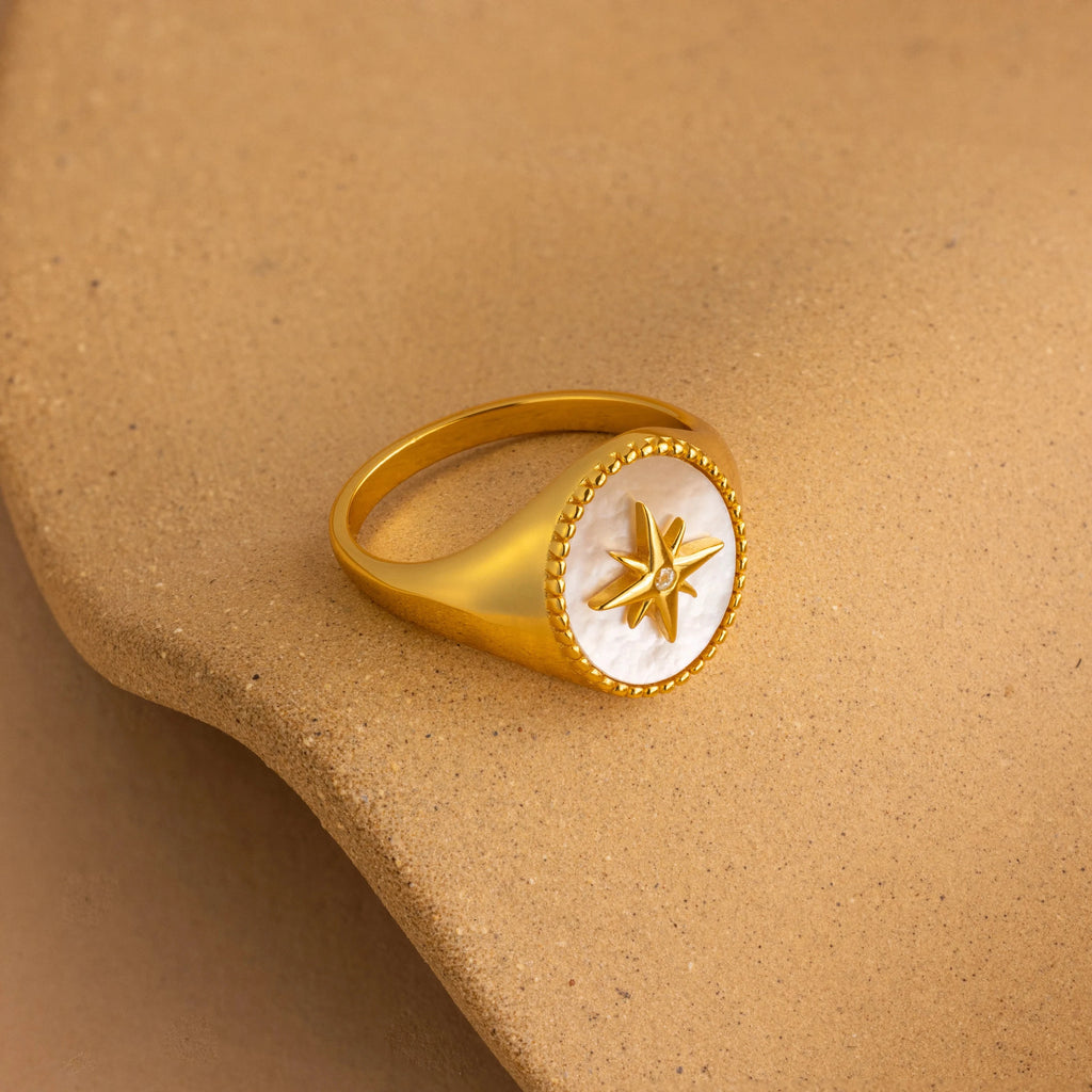 The North Star Signet Ring features a star design on a round white face and rests on a smooth beige surface—ideal statement jewelry.