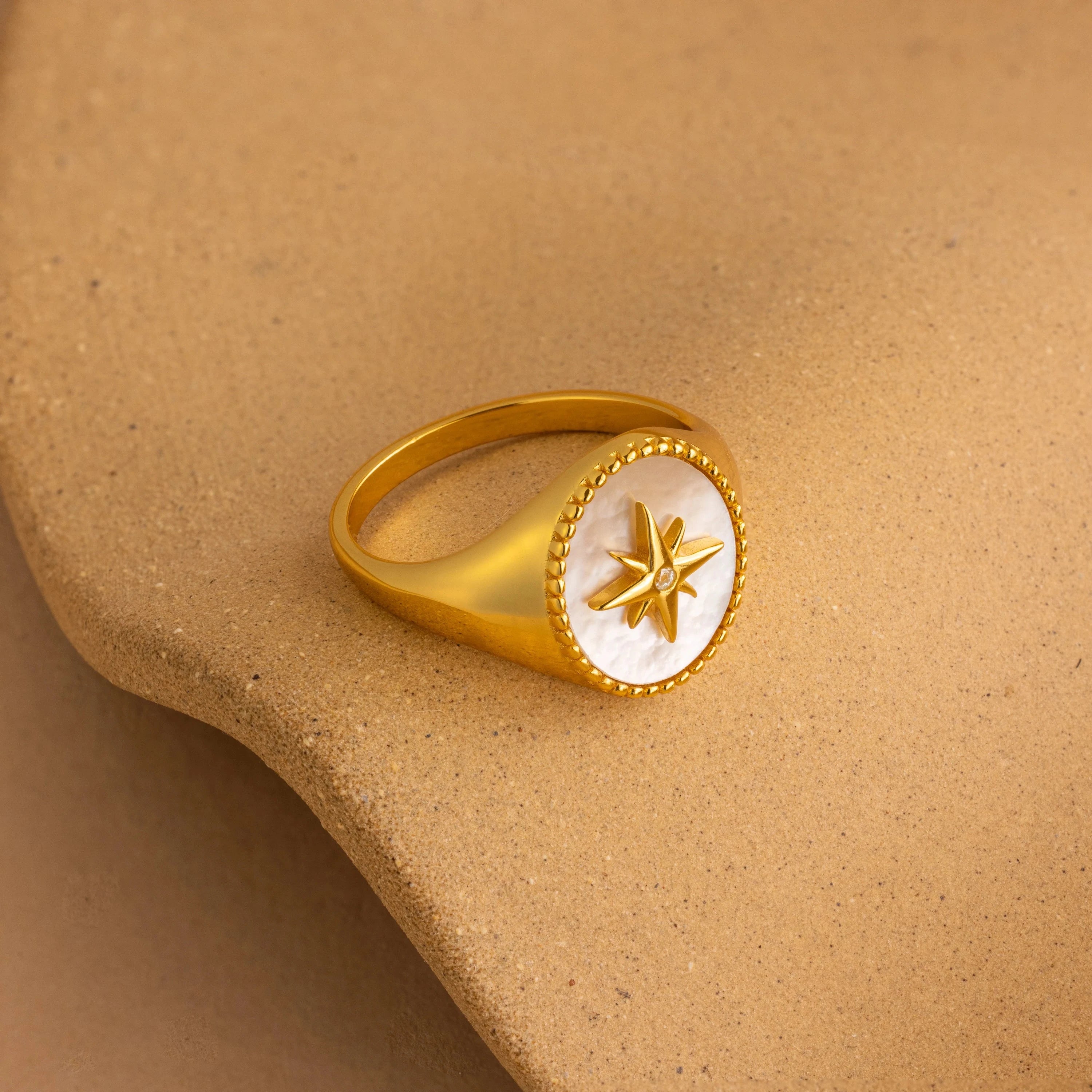 The North Star Signet Ring features a star design on a round white face and rests on a smooth beige surface—ideal statement jewelry.