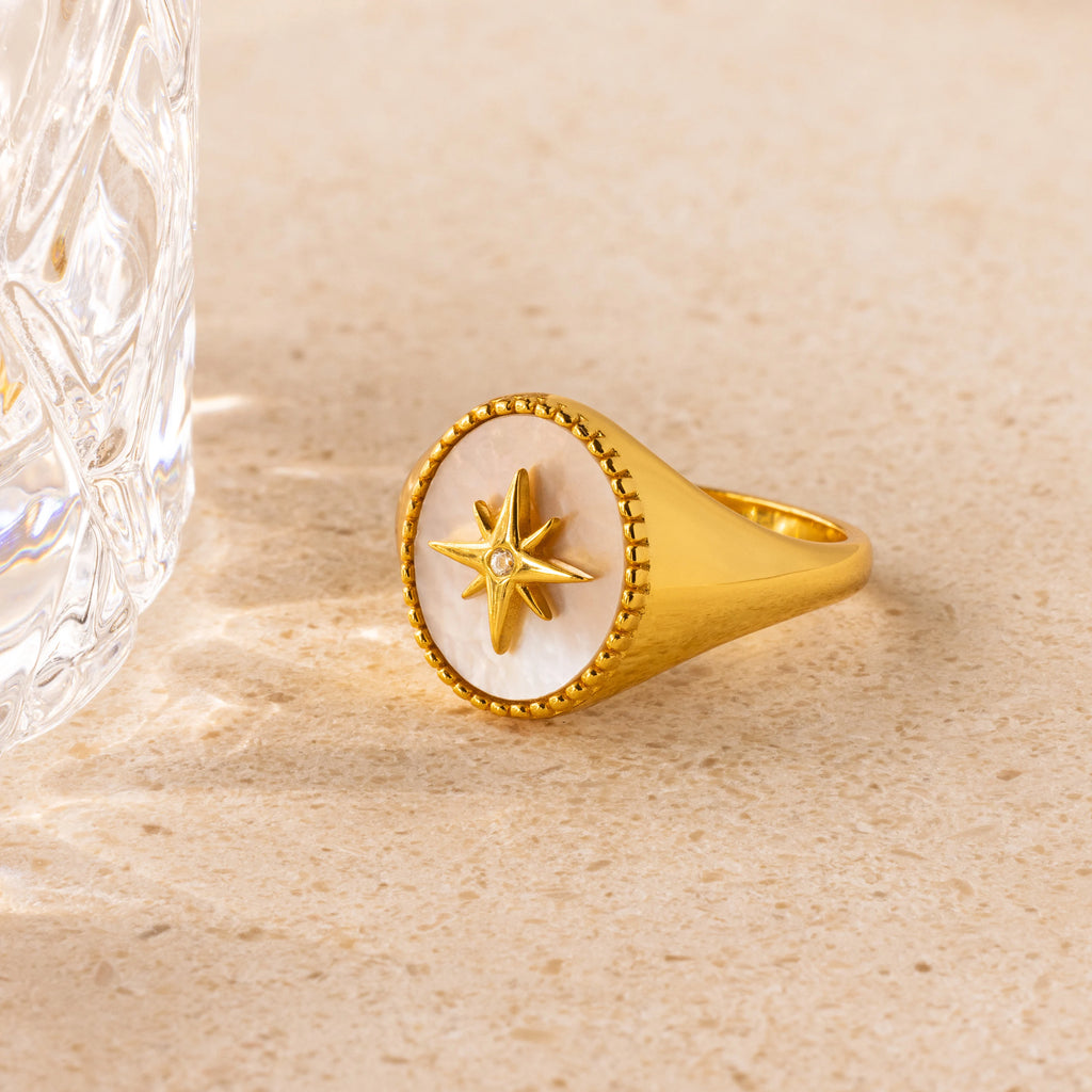 The North Star Signet Ring features a star design on a white inlay, displayed on a light stone surface beside a glass—a stunning piece for making a statement.