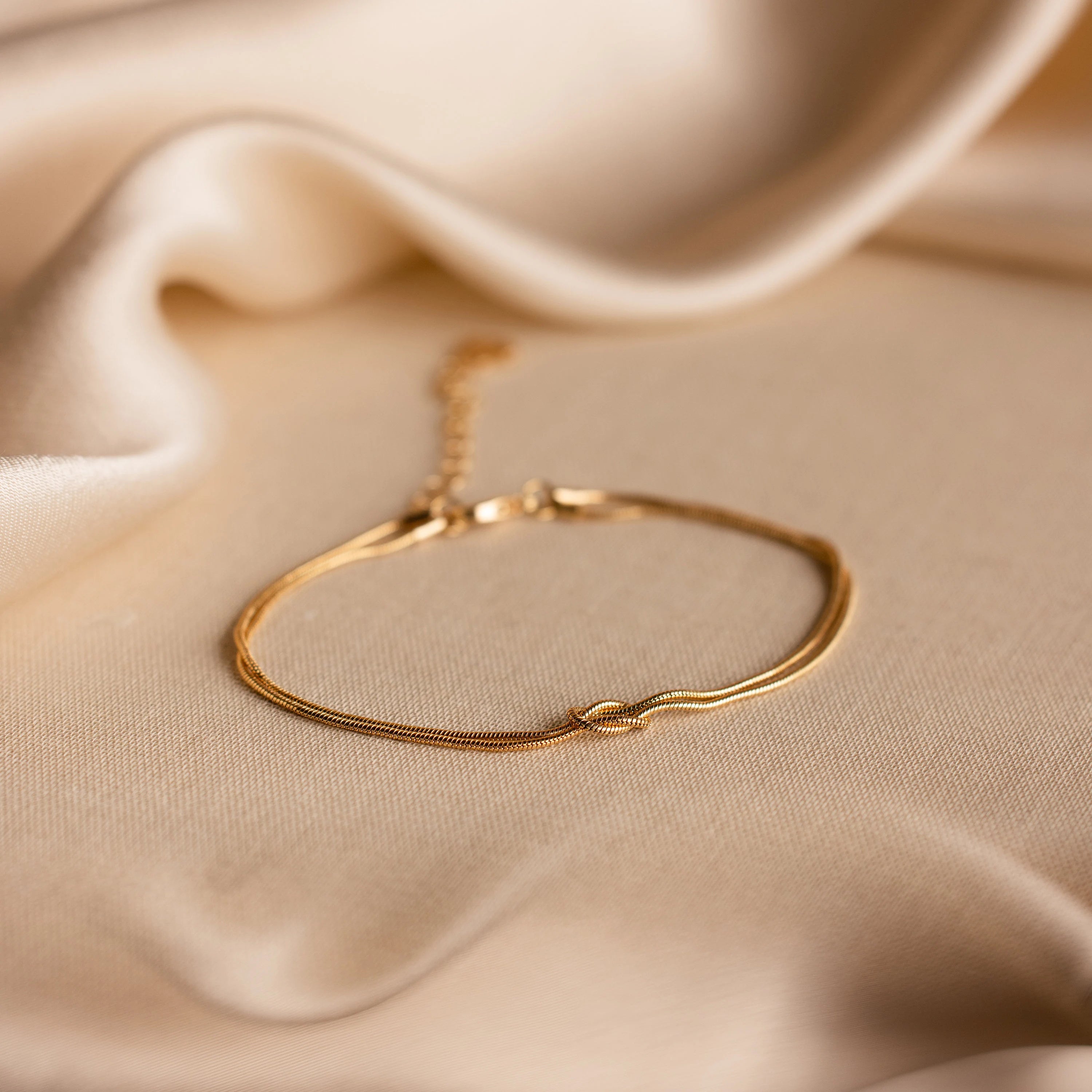 The Love Knot Bracelet features a delicate double chain with a subtle knot detail, beautifully displayed on soft beige fabric—a thoughtful gift for couples.