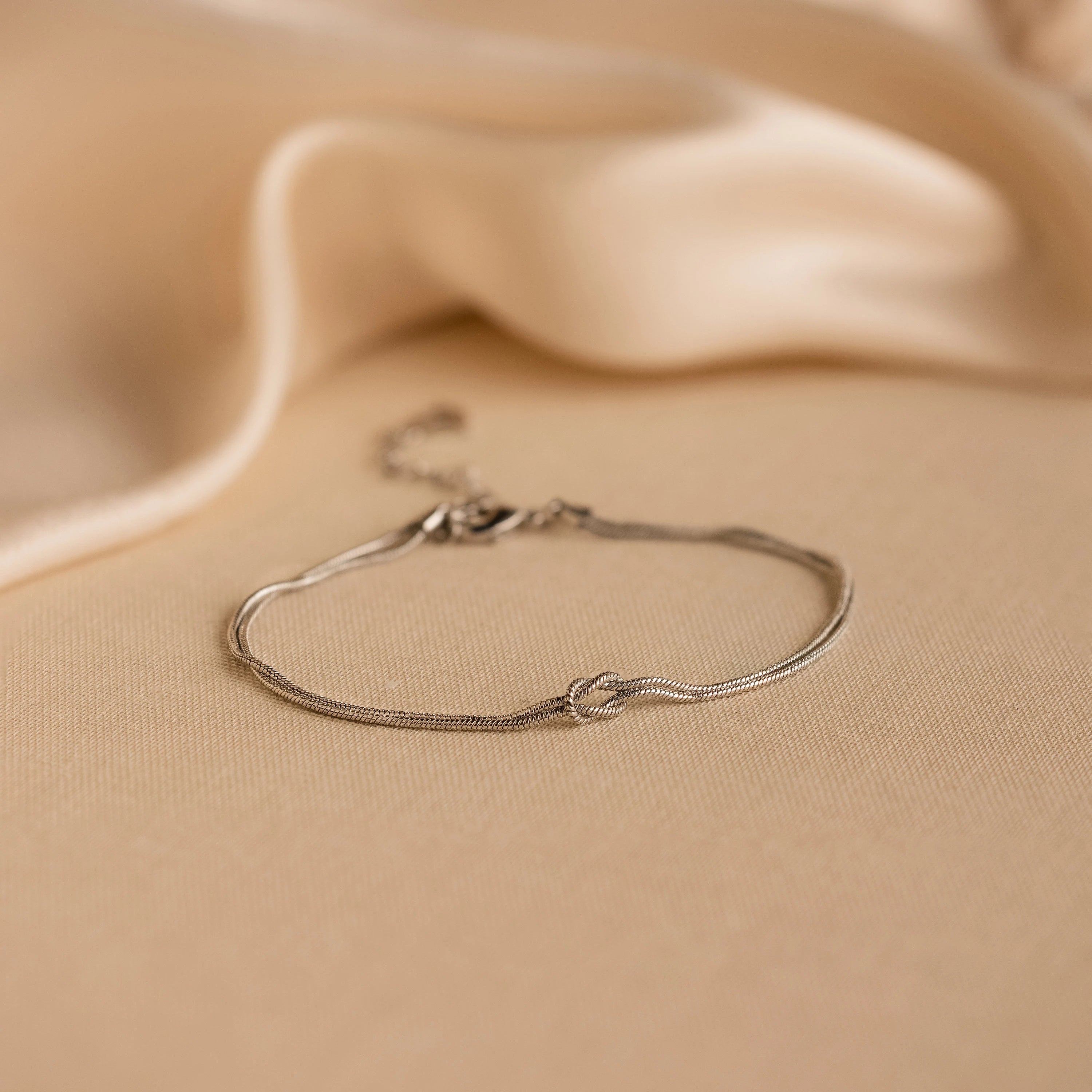 The Love Knot Bracelet features a delicate knot detail and double chain, elegantly displayed on soft beige fabric—an ideal sophisticated gift for couples.