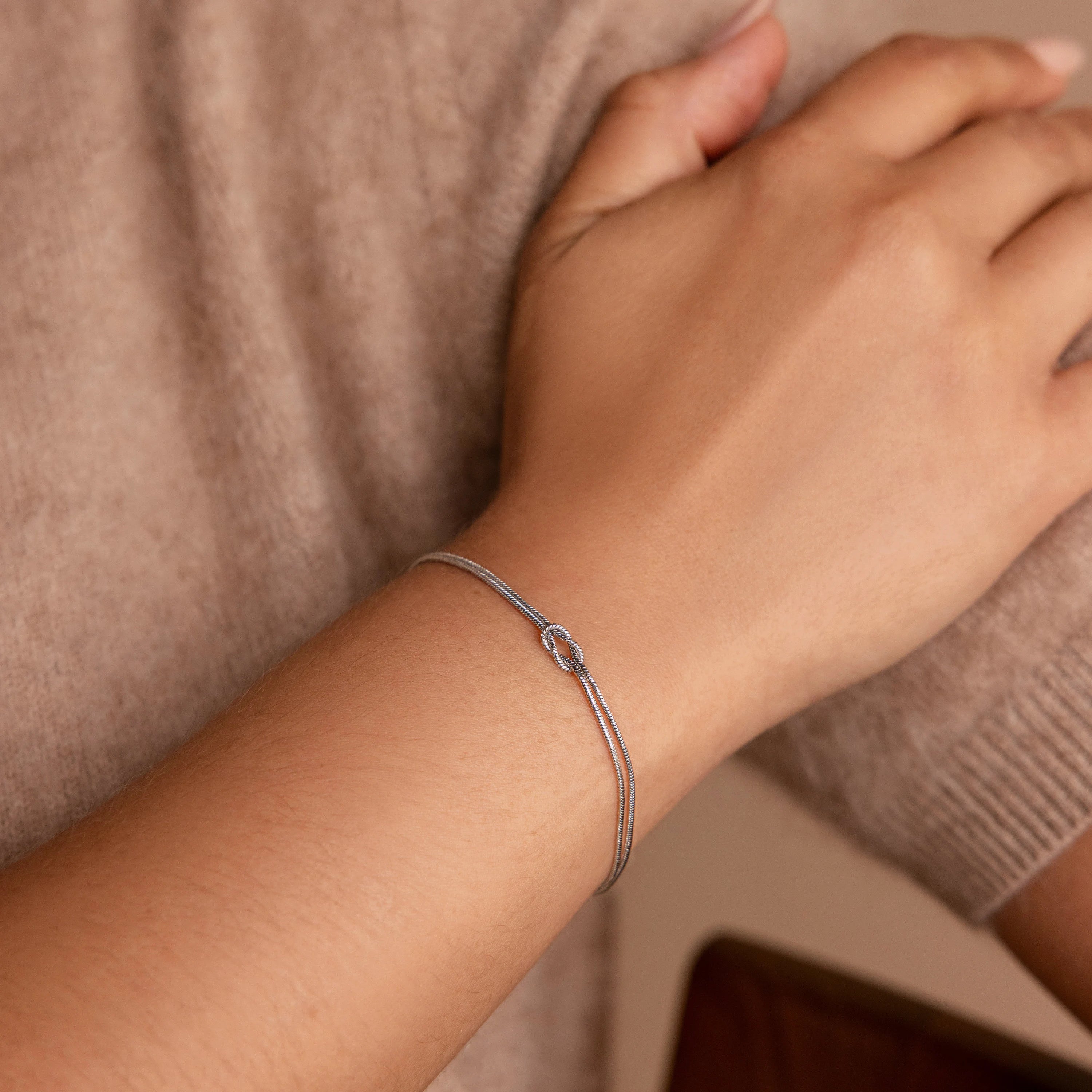 A hand wearing the Love Knot Bracelet in White Gold, featuring a delicate knot design and double chain against a beige sweater—a perfect gift for couples.
