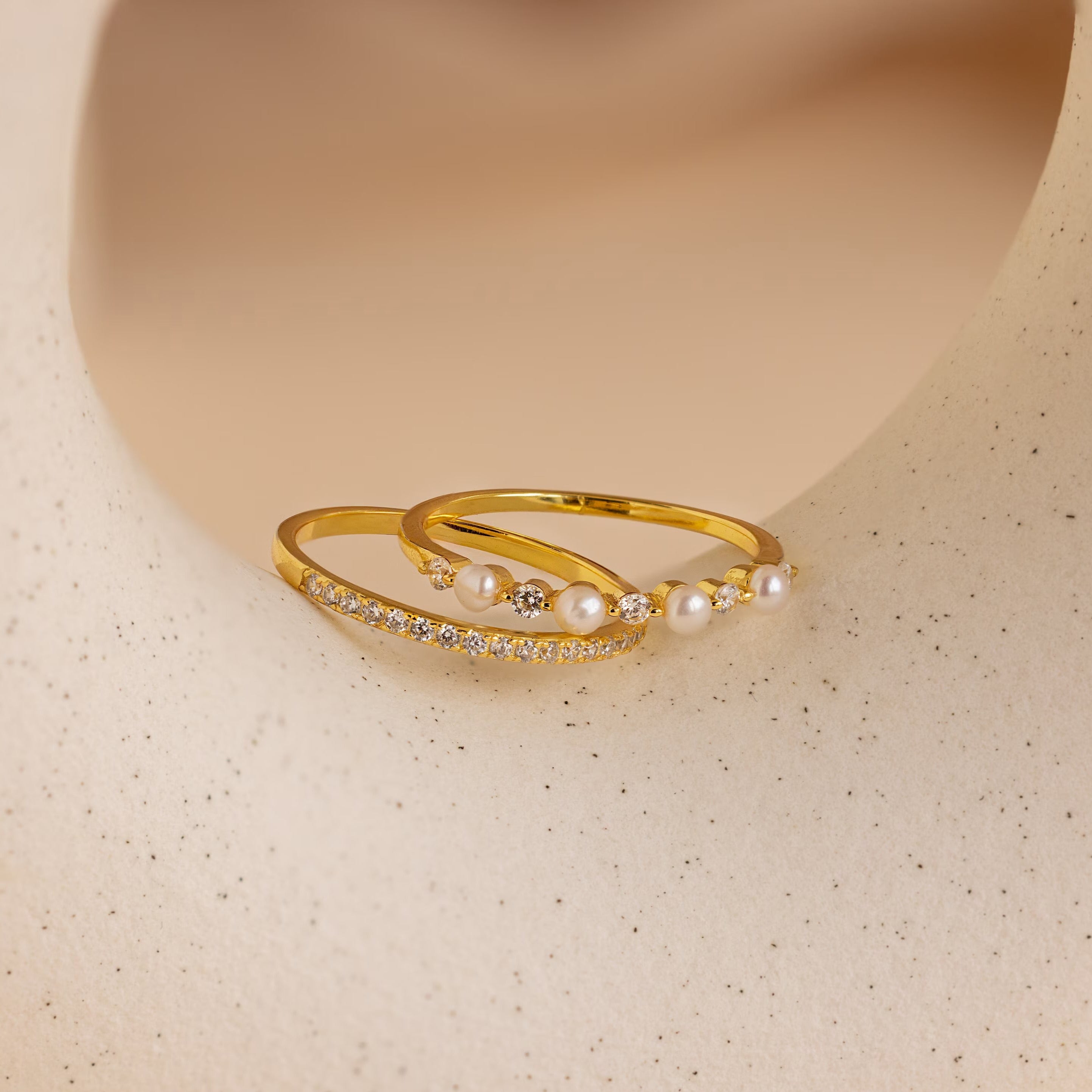 The Pave Pearl Stacking Ring Set features two delicate gold bands adorned with small diamonds and pearls, beautifully displayed on a smooth, cream-colored surface.