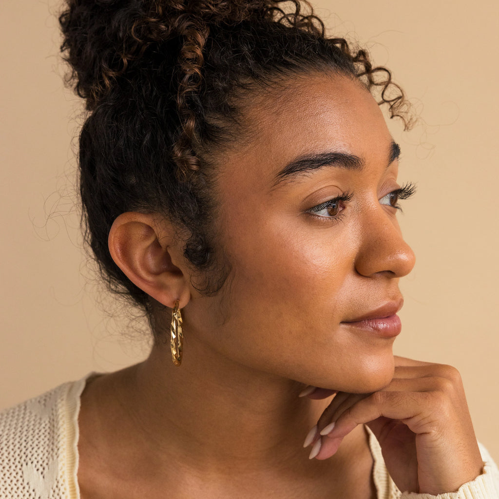 A woman with curly hair in a bun wears Large Twisted Hoops, posing with her hand under her chin and looking to the side—these bold earrings are the perfect summer accessory.