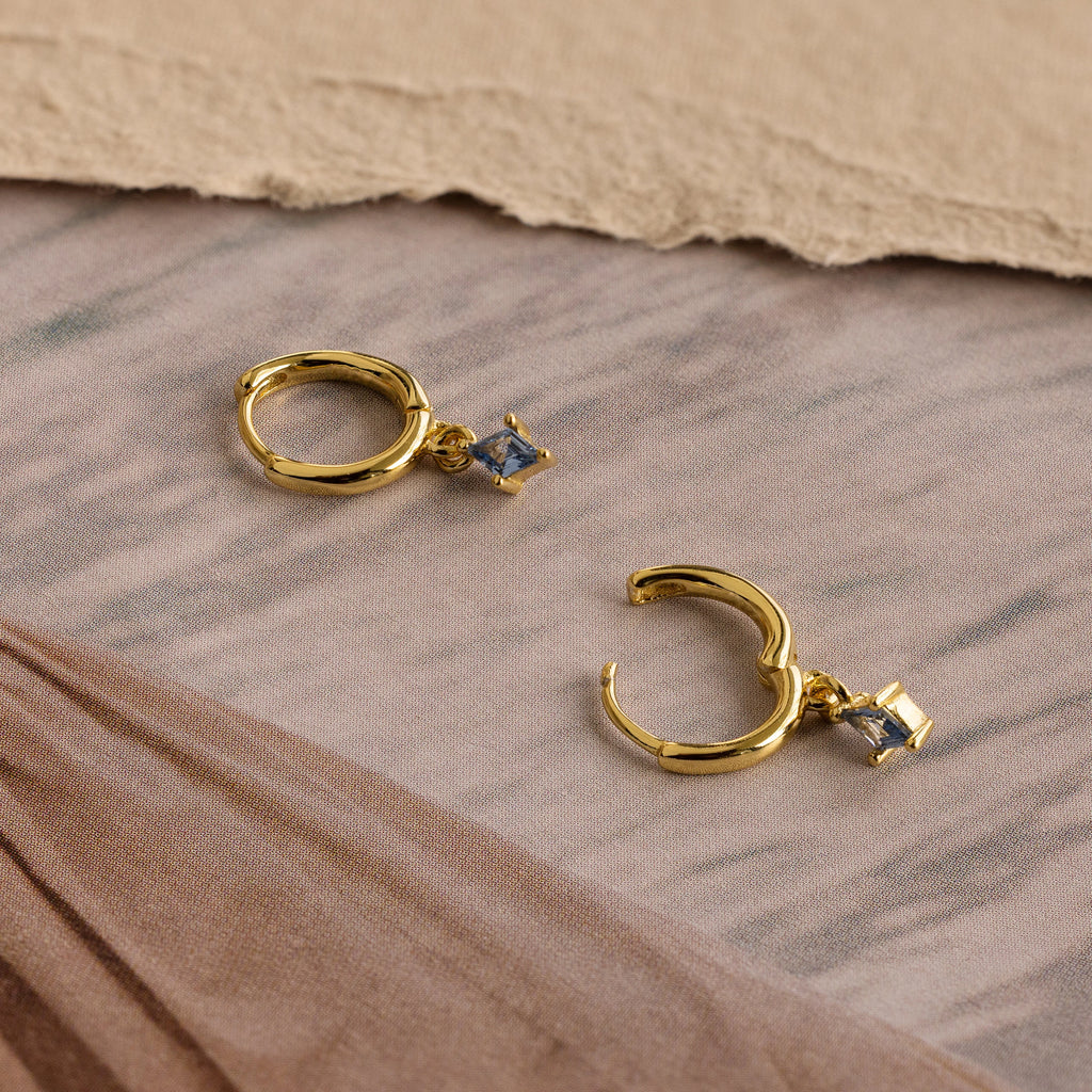 Birthstone Drop Huggies—gold huggie earrings with square blue gemstones, shown on a neutral fabric background. Ideal as birthstone earrings or personalized jewelry.