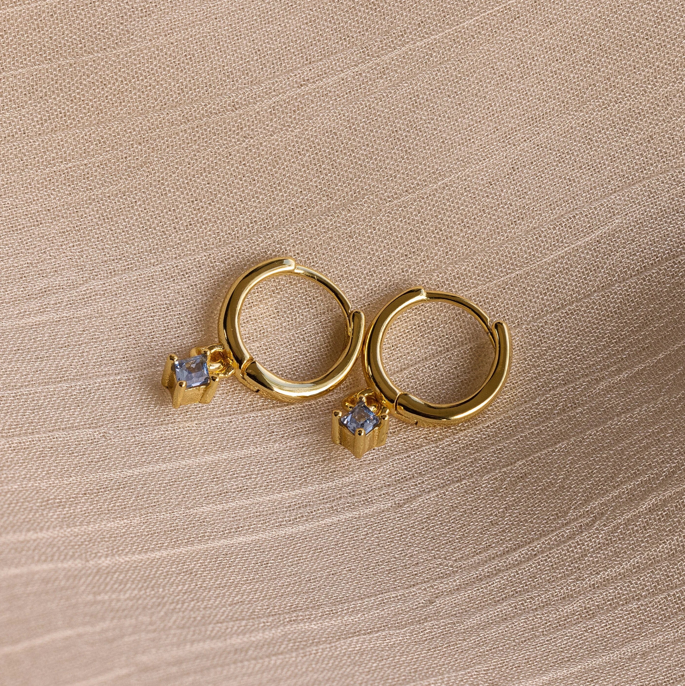 Birthstone Drop Huggies—gold huggie earrings featuring small square gemstone pendants, shown on textured beige fabric. Ideal as birthstone earrings or a meaningful piece of personalized jewelry.