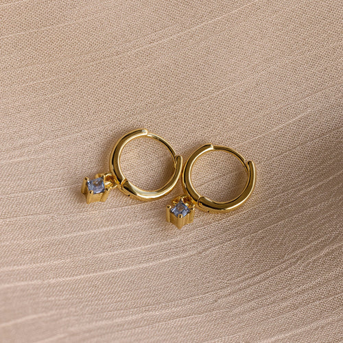 Birthstone Drop Huggies—gold huggie earrings featuring small square gemstone pendants, shown on textured beige fabric. Ideal as birthstone earrings or a meaningful piece of personalized jewelry.