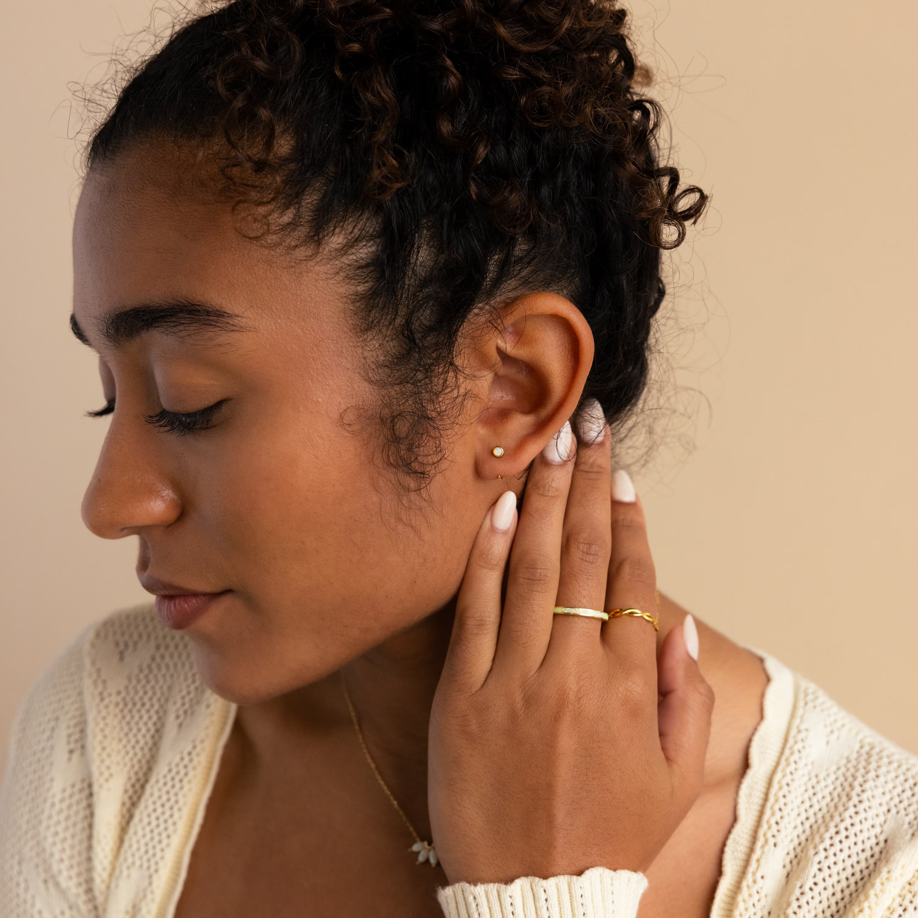 A woman with curly hair and patterned nails, eyes closed, touches her ear adorned with gold Opal Threader Huggies celestial jewelry.