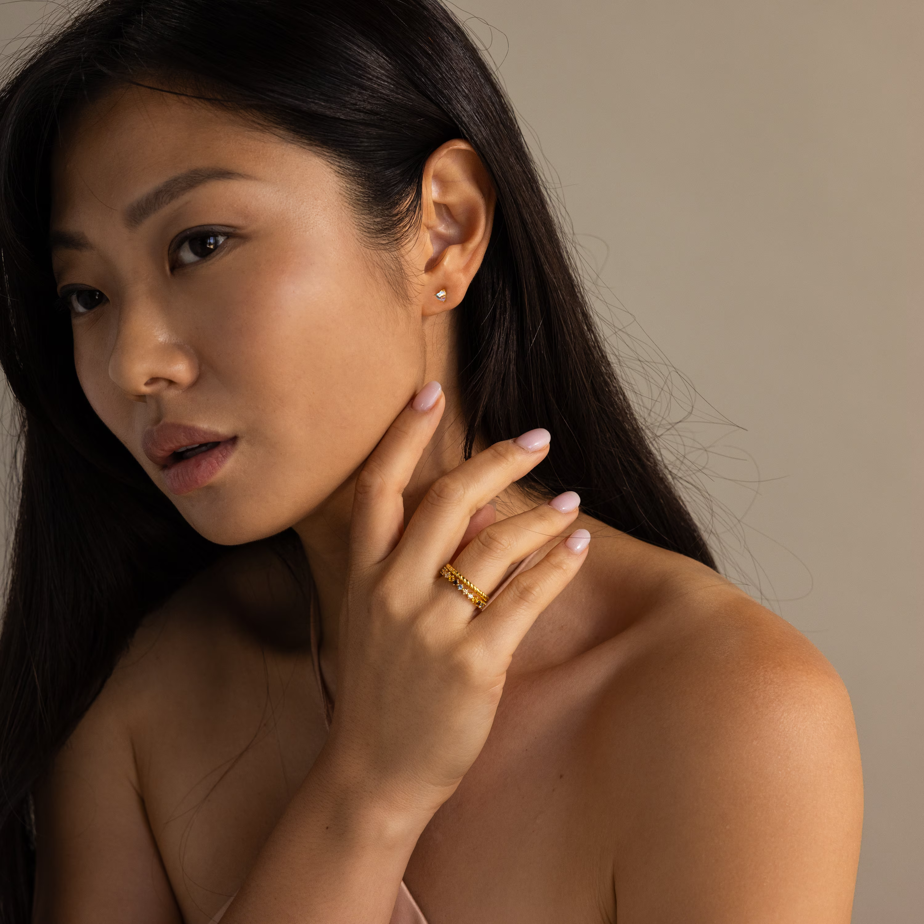 A woman with long dark hair showcases the Moonstone Heart Studs and a gold ring, gently touching her neck against a neutral background.