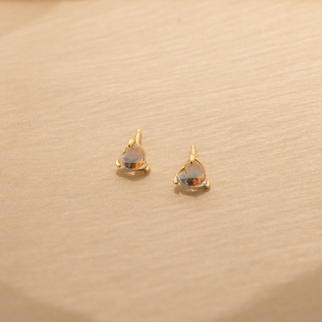 Moonstone Heart Studs: small gold stud earrings with heart-shaped iridescent moonstones, displayed on a beige textured surface.