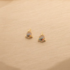 Moonstone Heart Studs: small gold stud earrings with heart-shaped iridescent moonstones, displayed on a beige textured surface.