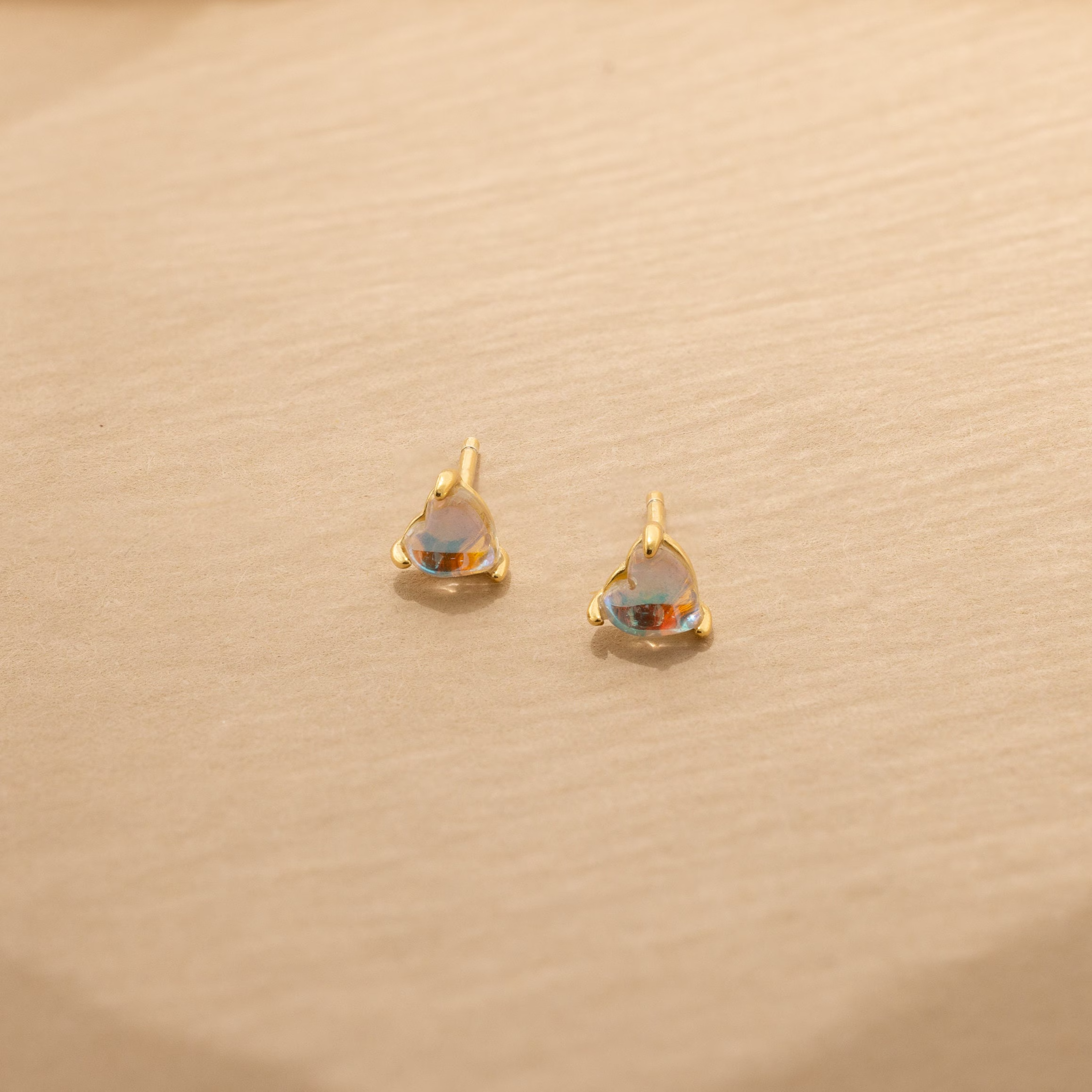 Moonstone Heart Studs: small gold stud earrings with heart-shaped iridescent moonstones, displayed on a beige textured surface.