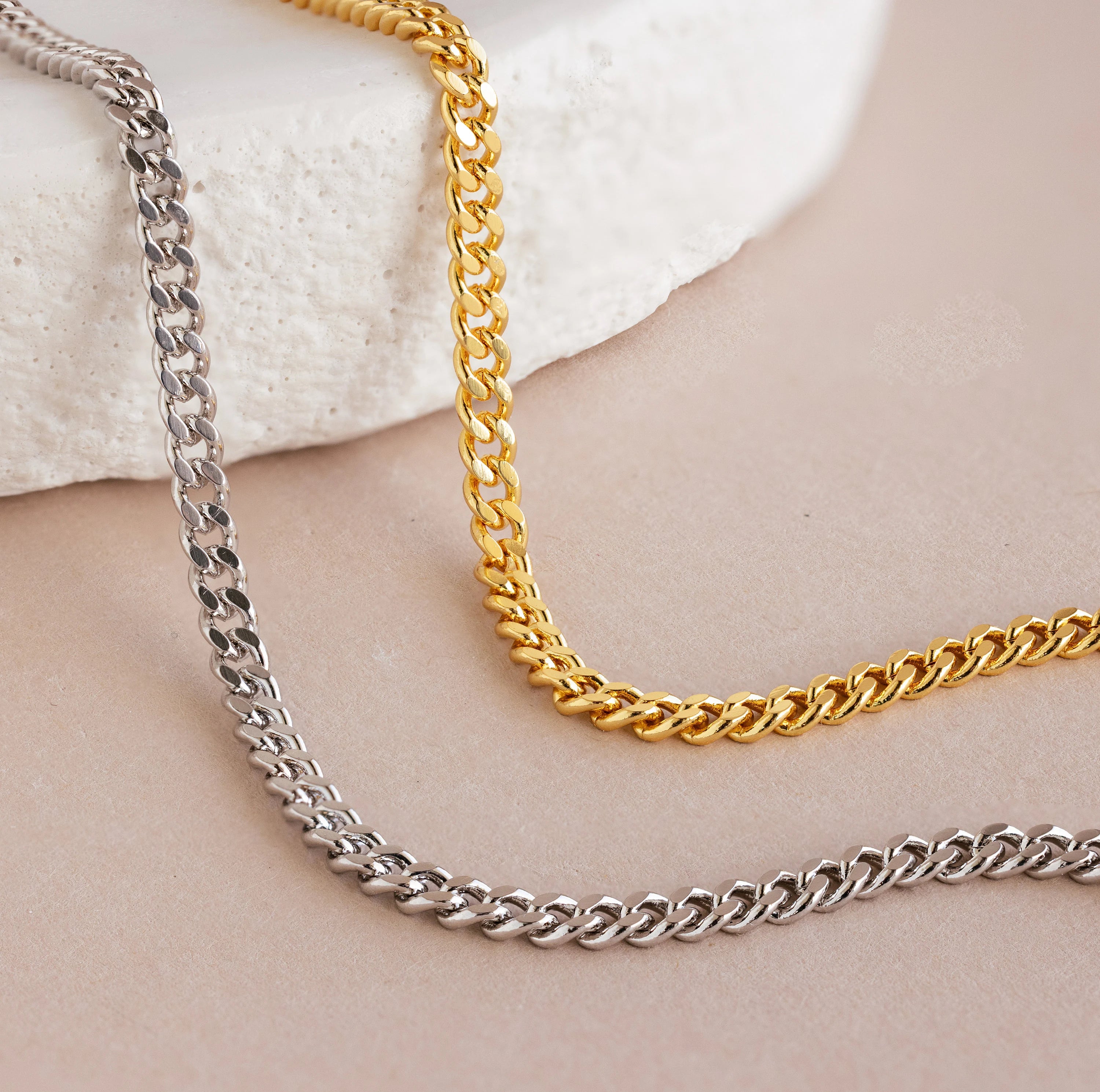 Gold and silver chain necklaces rest on a beige surface beside a white stone, offering a timeless, minimal look. Complete the style with the Curb Chain Bracelet for effortless elegance.