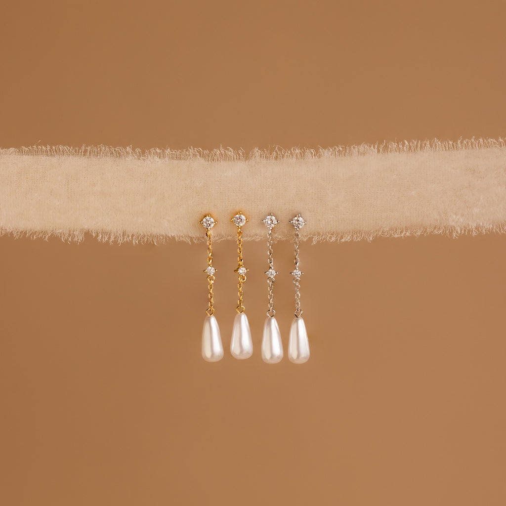 The Diamond Pearl Chain Earrings, featuring elegant drop pearl designs and a teardrop charm, hang on soft beige textured fabric with a brown background.