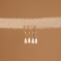 The Diamond Pearl Chain Earrings, featuring elegant drop pearl designs and a teardrop charm, hang on soft beige textured fabric with a brown background.