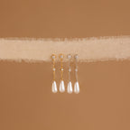 The Diamond Pearl Chain Earrings, featuring elegant drop pearl designs and a teardrop charm, hang on soft beige textured fabric with a brown background.