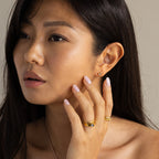 A woman with long dark hair touches her face, looking thoughtful as she wears gold rings and delicate Moonstone Studs.