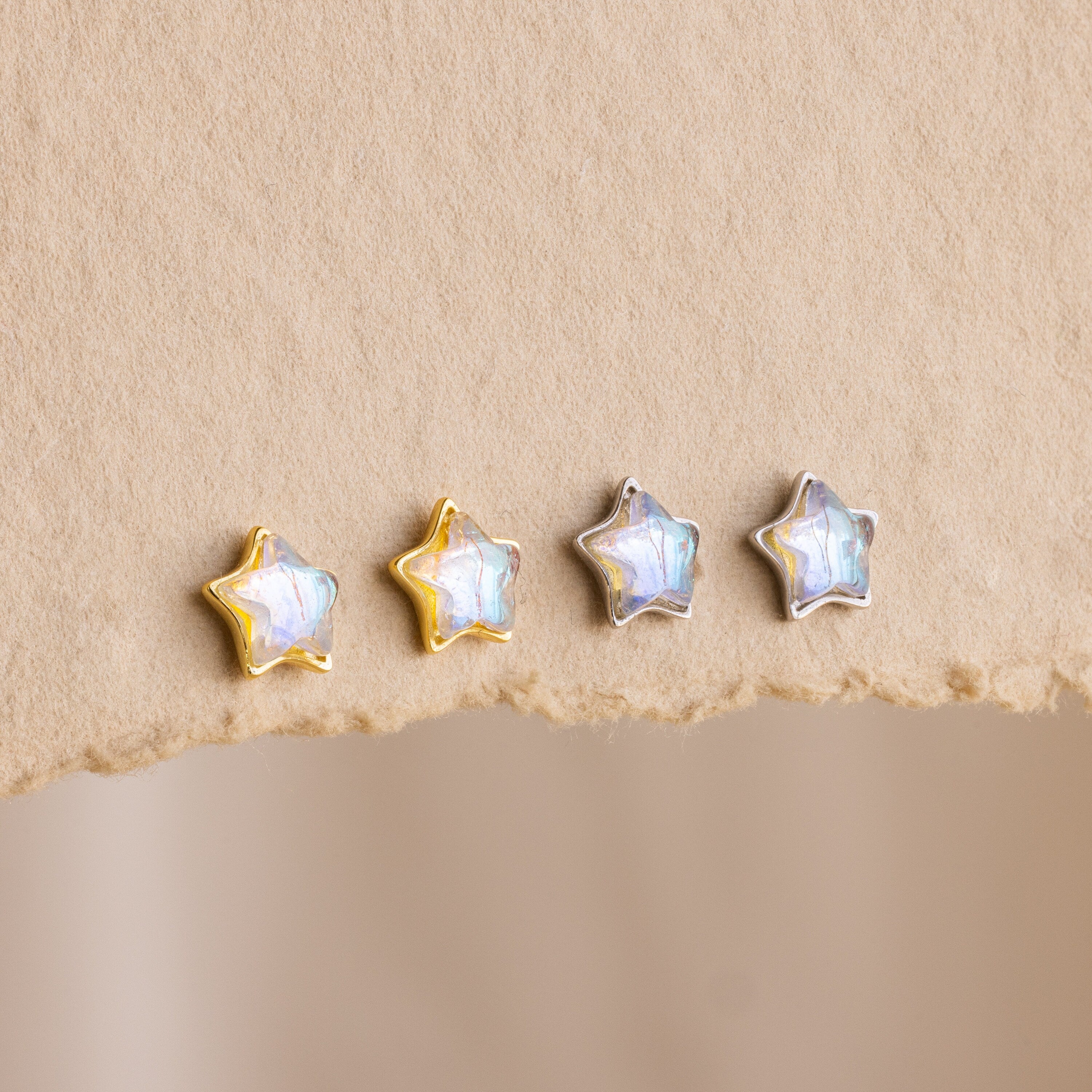 Four Moonstone Star Studs—two with gold edges and two with silver—rest on a beige textured surface, perfect for adding a celestial touch to your style.