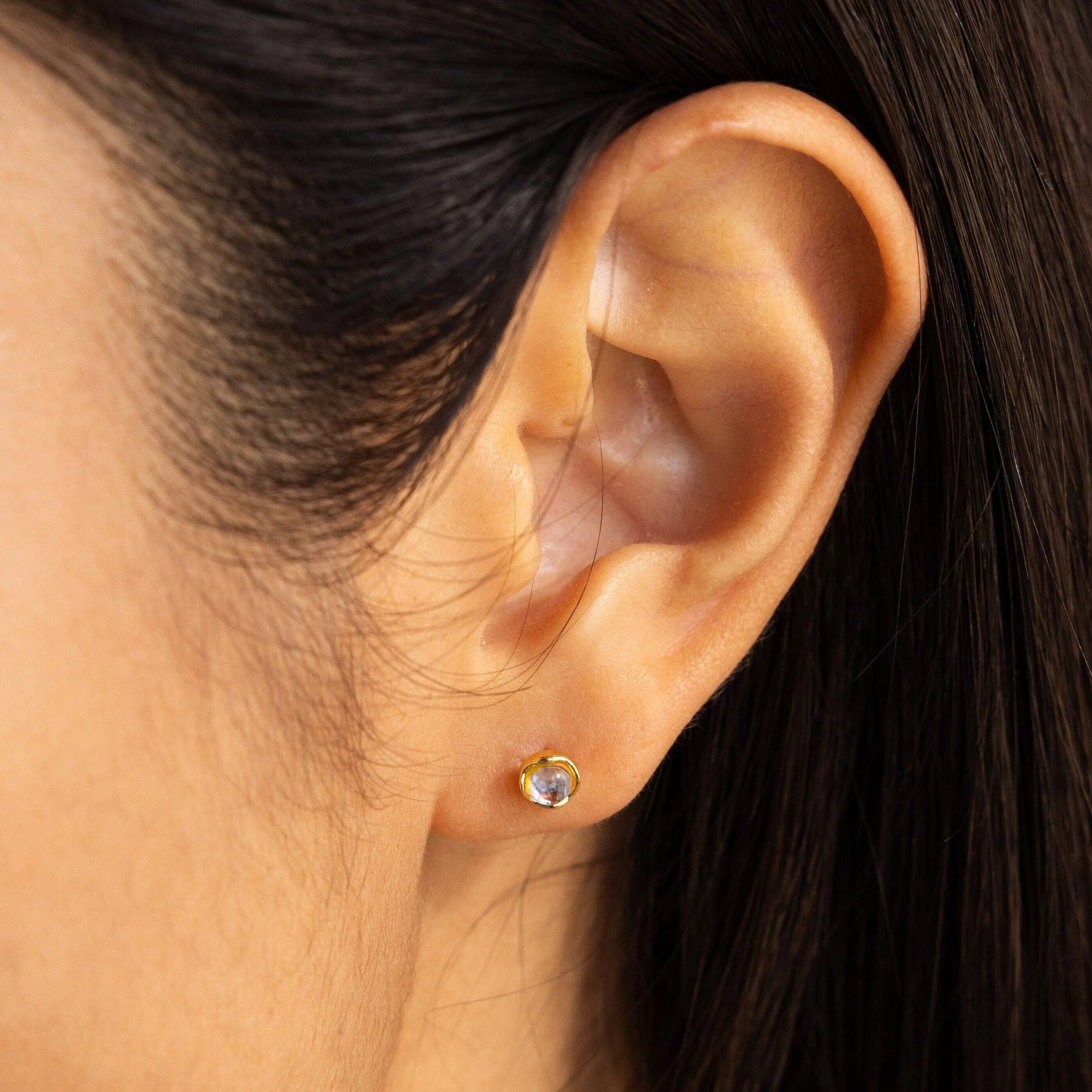 A close-up of an ear wearing Moonstone Studs—small gold earrings with clear gemstones—showcasing their subtle, celestial beauty and luminous charm.