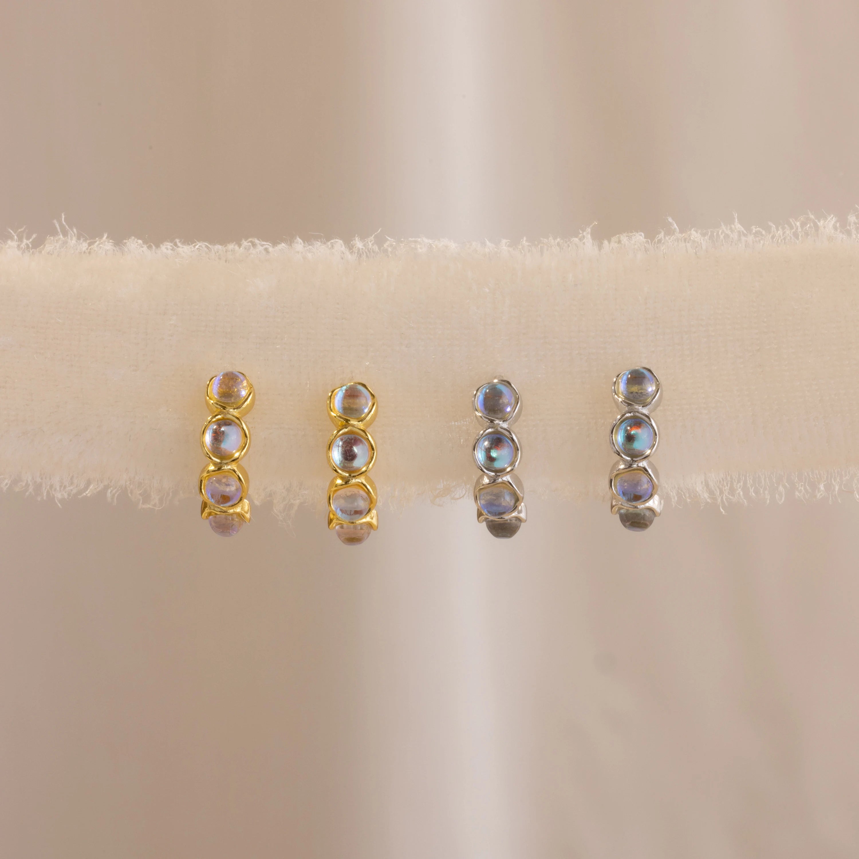 Four pairs of elegant earrings—two gold, two silver studs—are displayed on a soft beige fabric background, perfect for pairing with the Clear Moonstone Huggies.