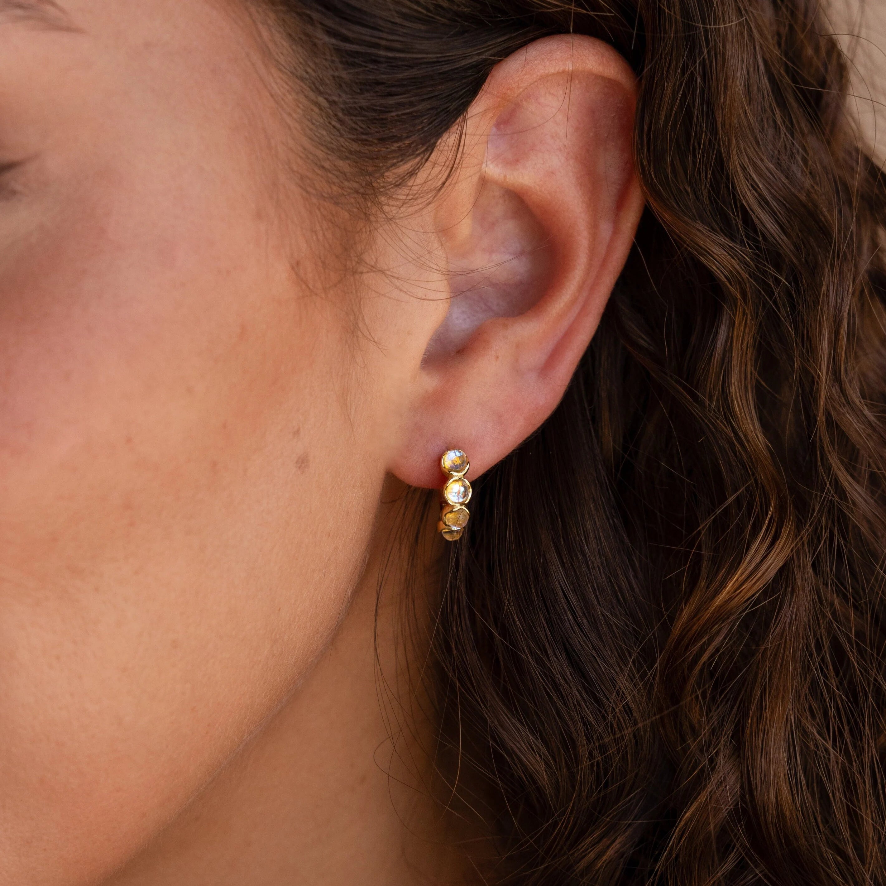 Close-up of a woman's ear adorned with Clear Moonstone Huggies, featuring three small round moonstones and wavy brown hair in view.