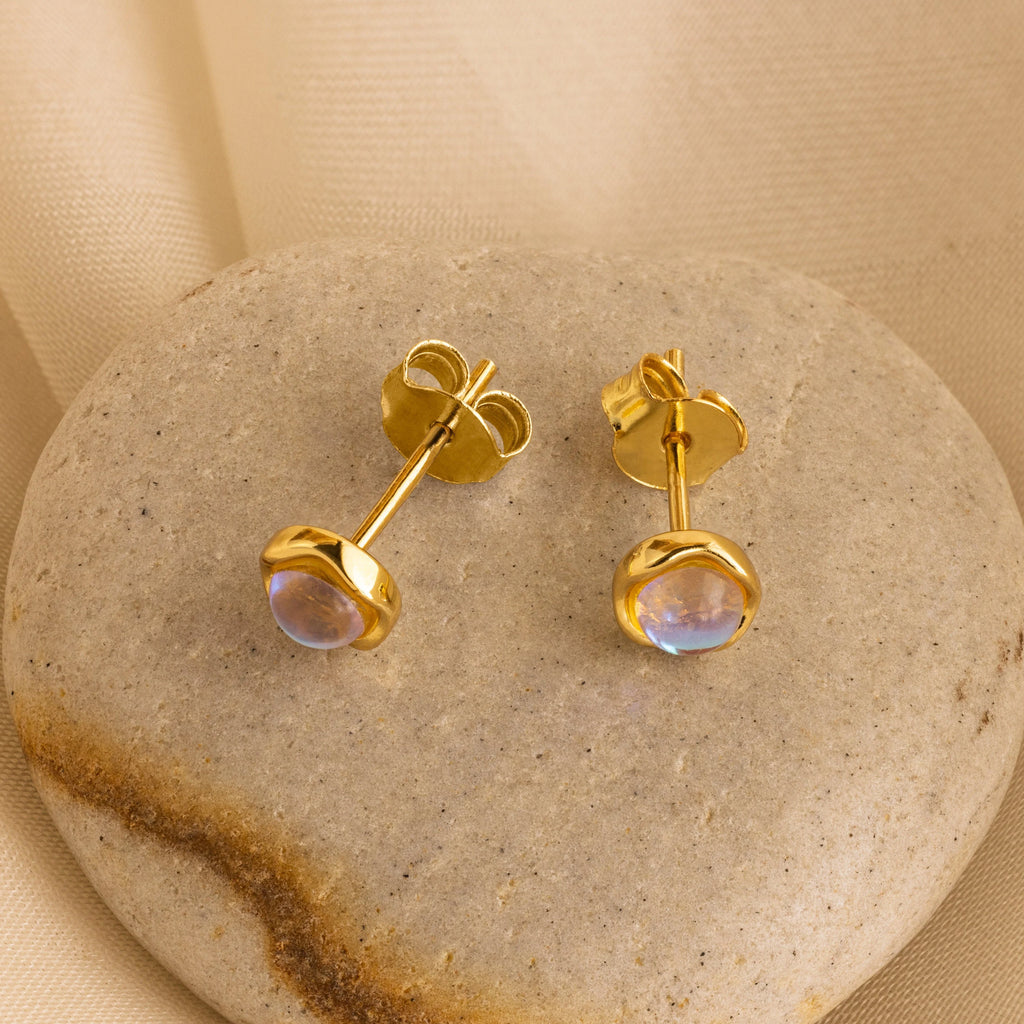 Moonstone Studs: Gold stud earrings with round moonstones, elegantly displayed on a smooth stone against a beige fabric background—ideal for celestial jewelry lovers.