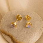 Moonstone Studs: Gold stud earrings with round moonstones, elegantly displayed on a smooth stone against a beige fabric background—ideal for celestial jewelry lovers.