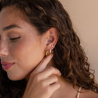 A woman with curly hair showcases several elegant earrings, including Clear Moonstone Huggies and diamond styles, as she softly touches her earlobe.