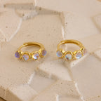 Two elegant Clear Moonstone Huggies, each gold hoop earring adorned with three clear moonstone gemstones, are displayed on a textured beige surface.