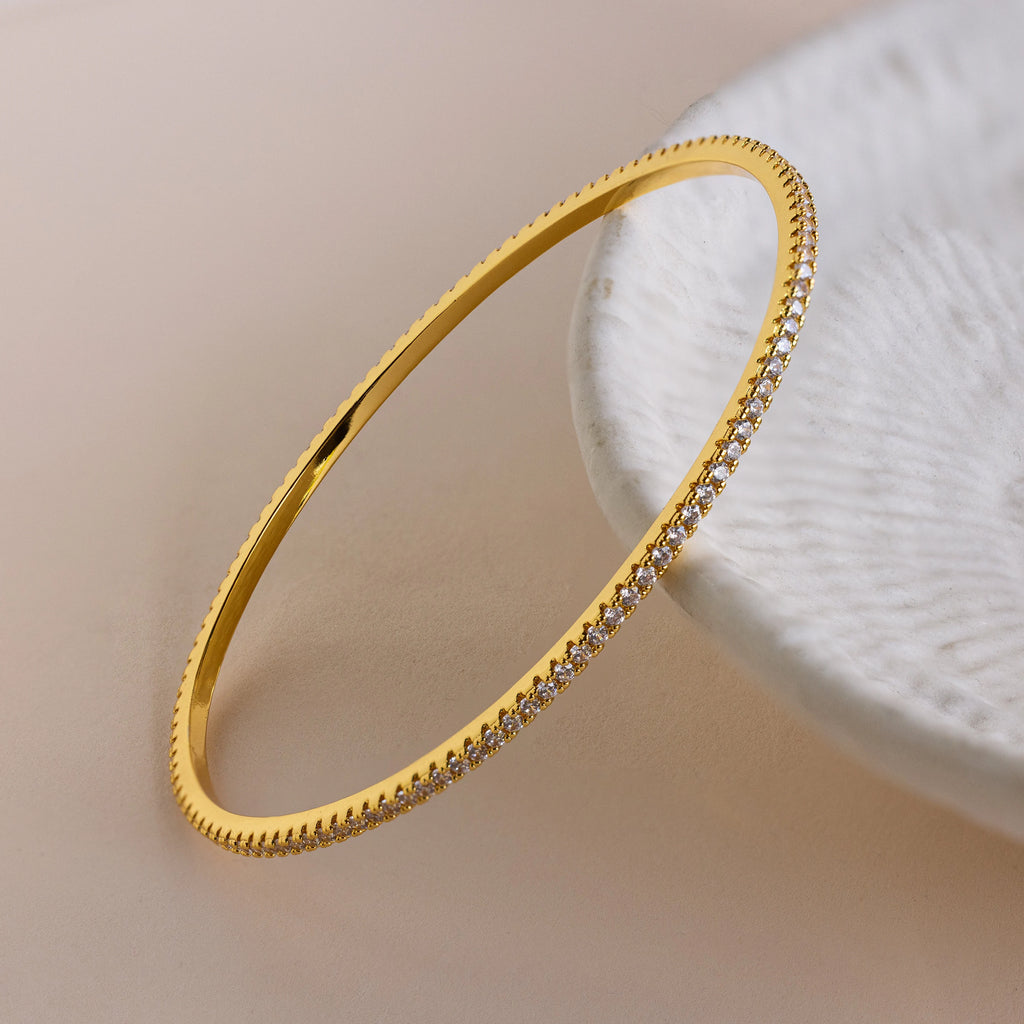 The Classic Diamond Tennis Bangle, adorned with pavé stones, sparkles brilliantly on a white textured surface, adding radiant elegance to the timeless gold bracelet.