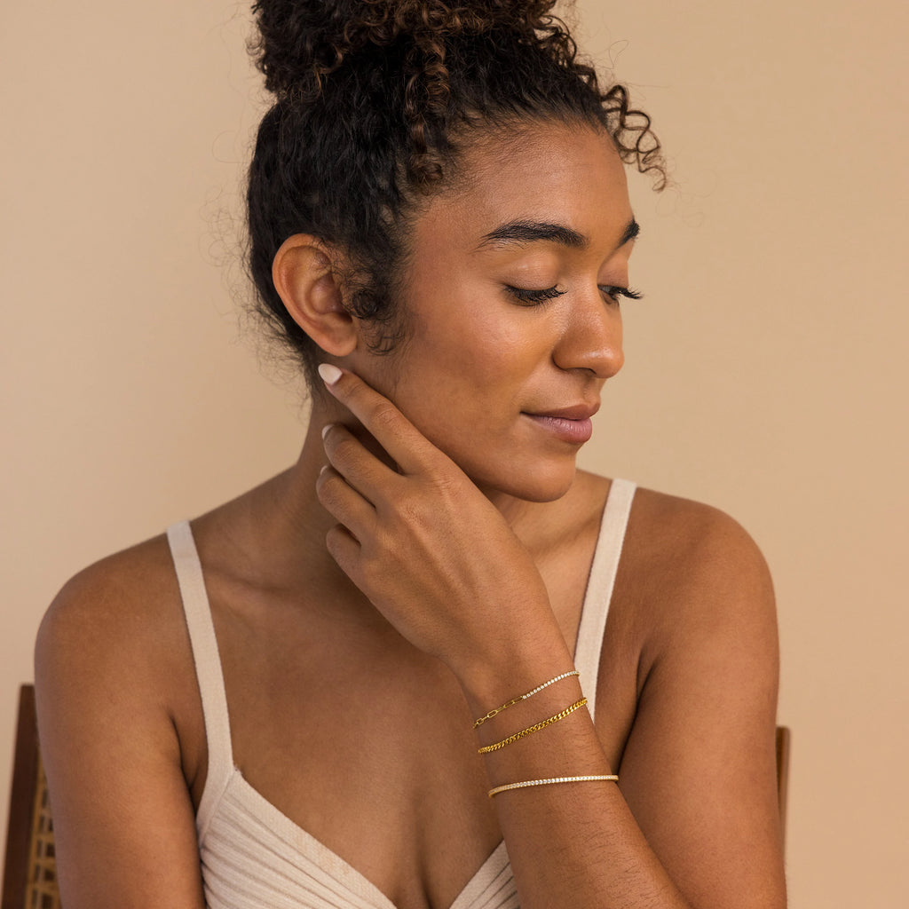 Woman with curly hair in a bun, wearing a beige top and the Classic Diamond Tennis Bangle, looks down softly.