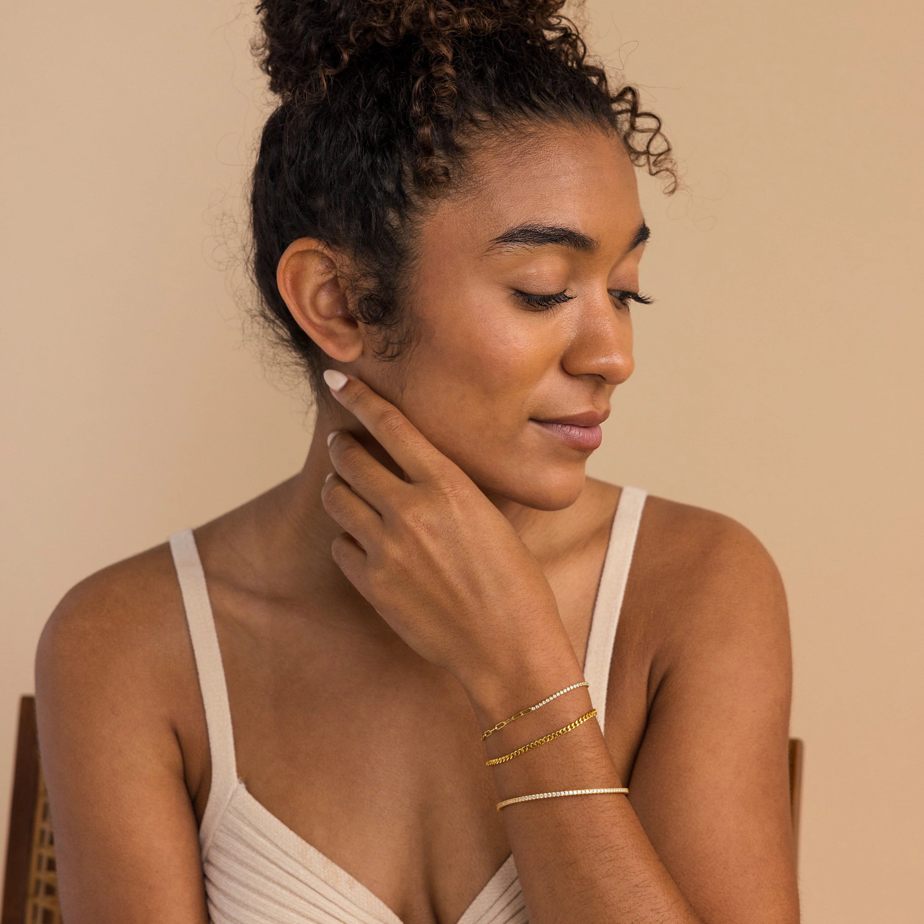 Woman with curly hair in a bun, wearing a beige top and the Classic Diamond Tennis Bangle, looks down softly.