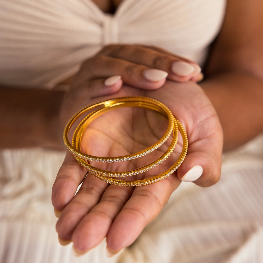 A person holds three gold bracelets, including the Classic Diamond Tennis Bangle, while wearing a cream pleated dress and nude nail polish.