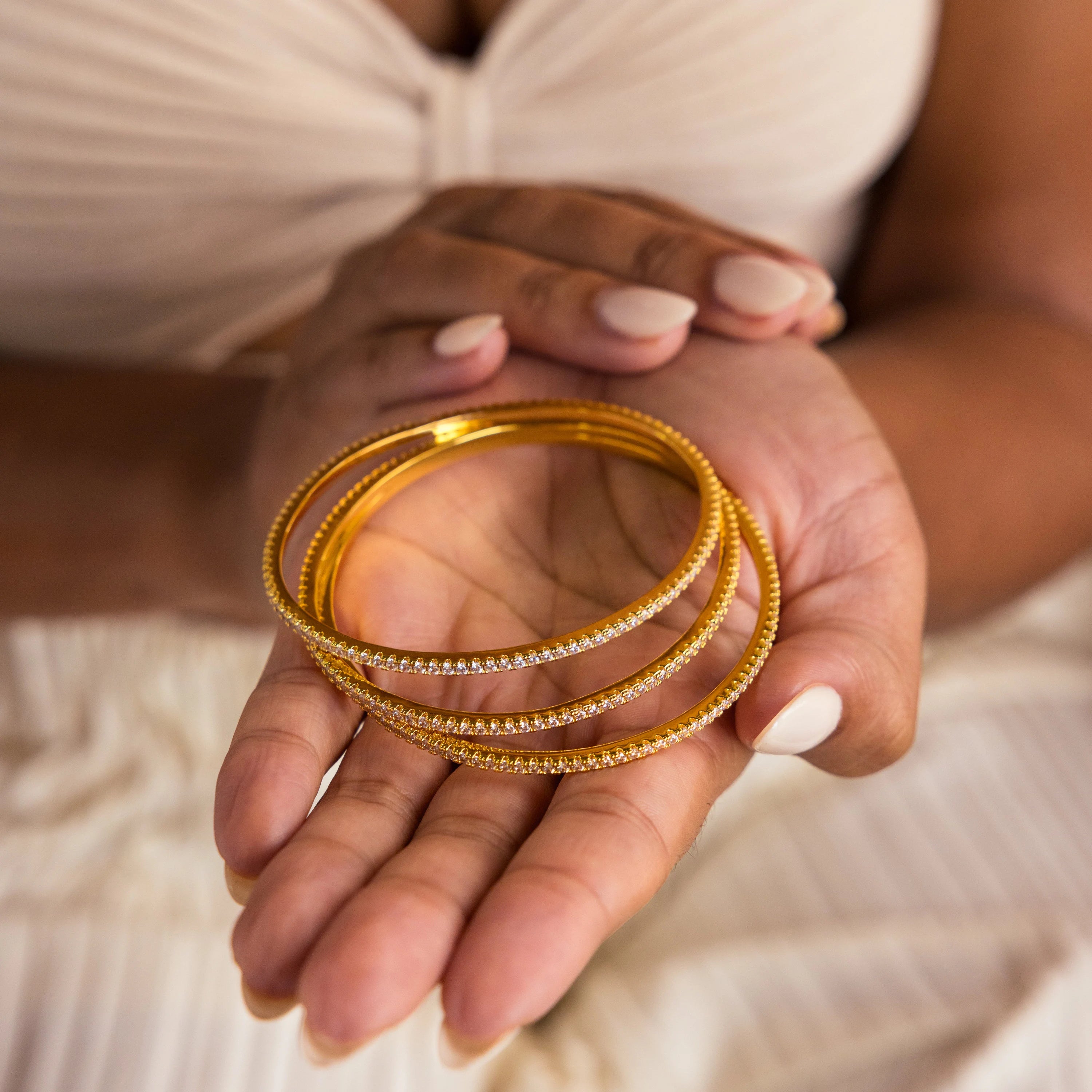 A person holds three gold bracelets, including the Classic Diamond Tennis Bangle, while wearing a cream pleated dress and nude nail polish.