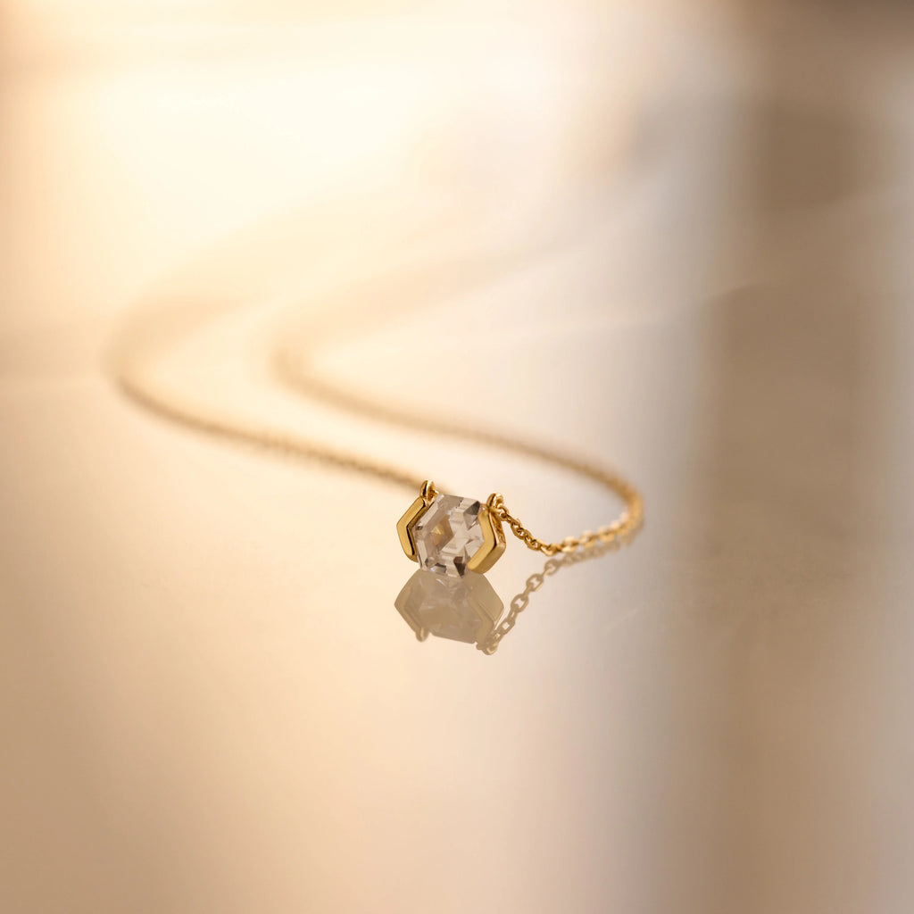 The Encased Hexagon Diamond Necklace, featuring a single diamond in a half-bezel setting, rests on a smooth, reflective surface and exudes modern sophistication.