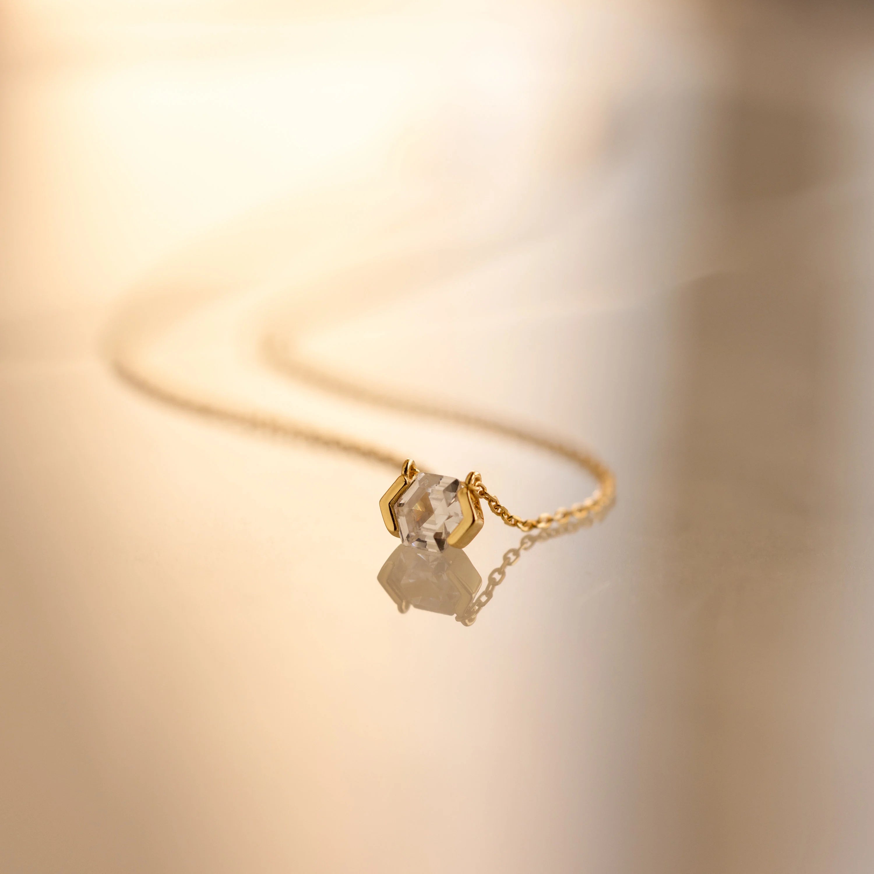 The Encased Hexagon Diamond Necklace, featuring a single diamond in a half-bezel setting, rests on a smooth, reflective surface and exudes modern sophistication.