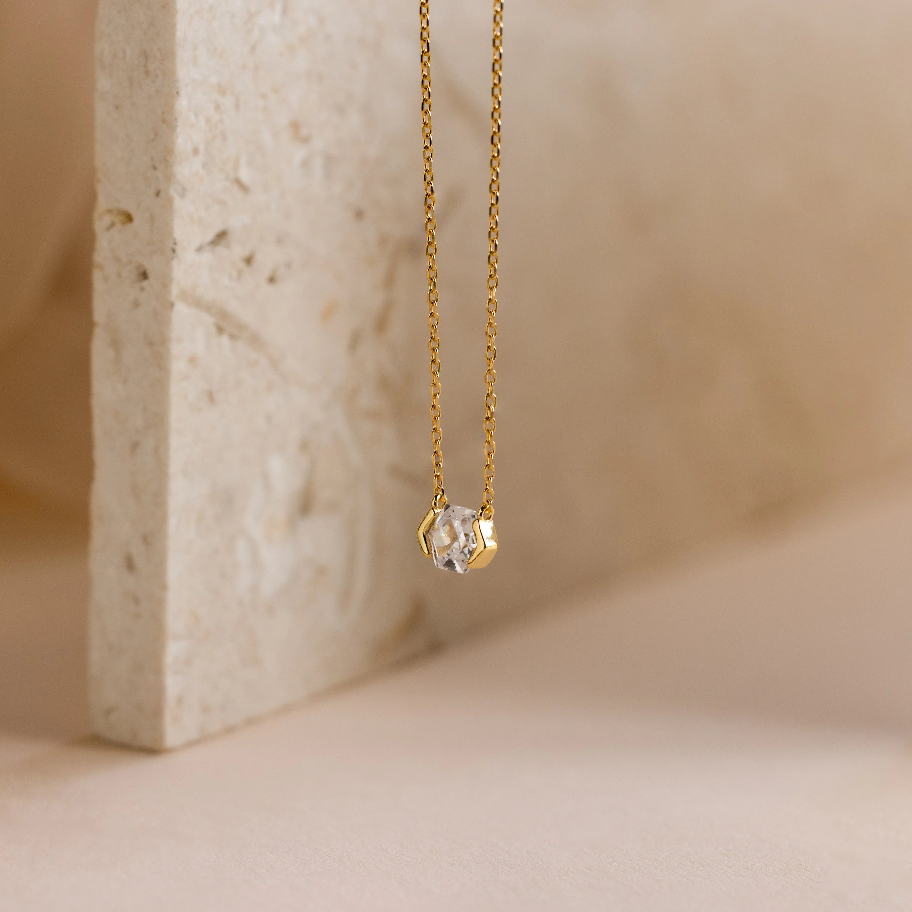 Encased Hexagon Diamond Necklace: A gold necklace with a clear hexagon diamond pendant in a sleek half-bezel setting, shown on textured stone—a modern, elegant accessory.