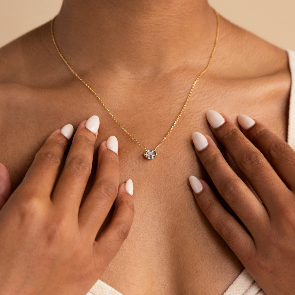 Close-up of a woman wearing the Encased Hexagon Diamond Necklace, featuring a single diamond pendant in a gold half-bezel hexagon setting, hands near her collarbone, capturing modern sophistication.