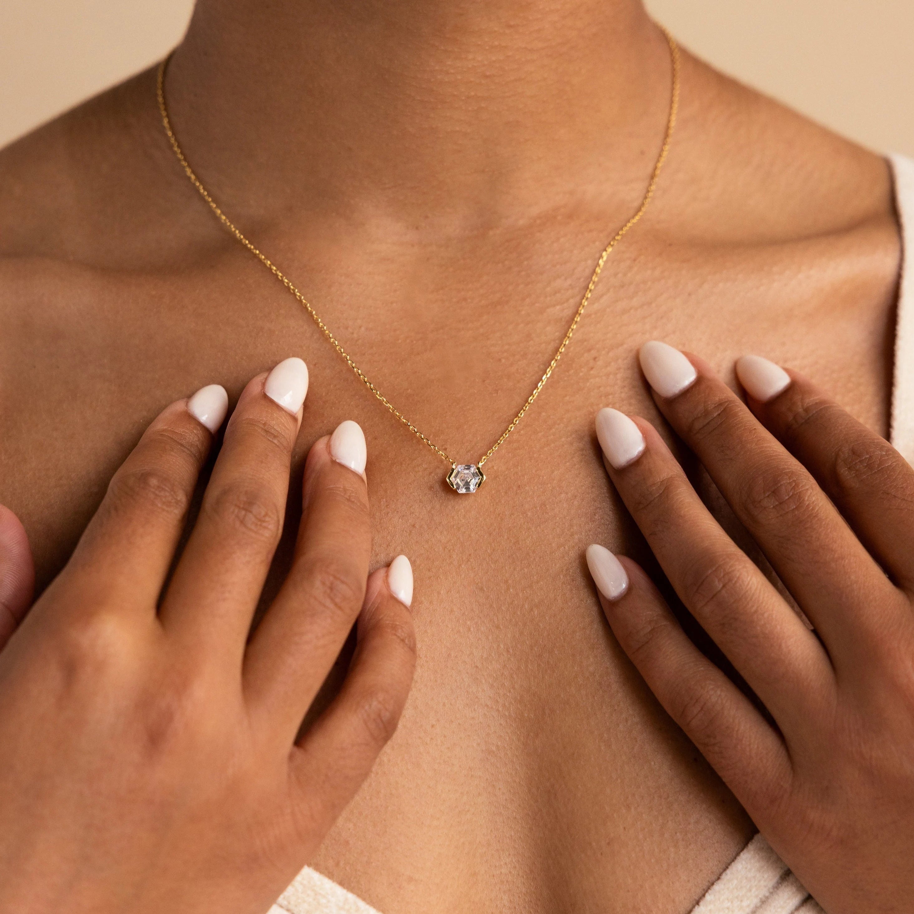 Close-up of a woman wearing the Encased Hexagon Diamond Necklace, featuring a single diamond pendant in a gold half-bezel hexagon setting, hands near her collarbone, capturing modern sophistication.