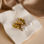 Baroque Pearl Earrings Set—gold hoops with dangling white pearls—rests elegantly on cream-colored fabric.