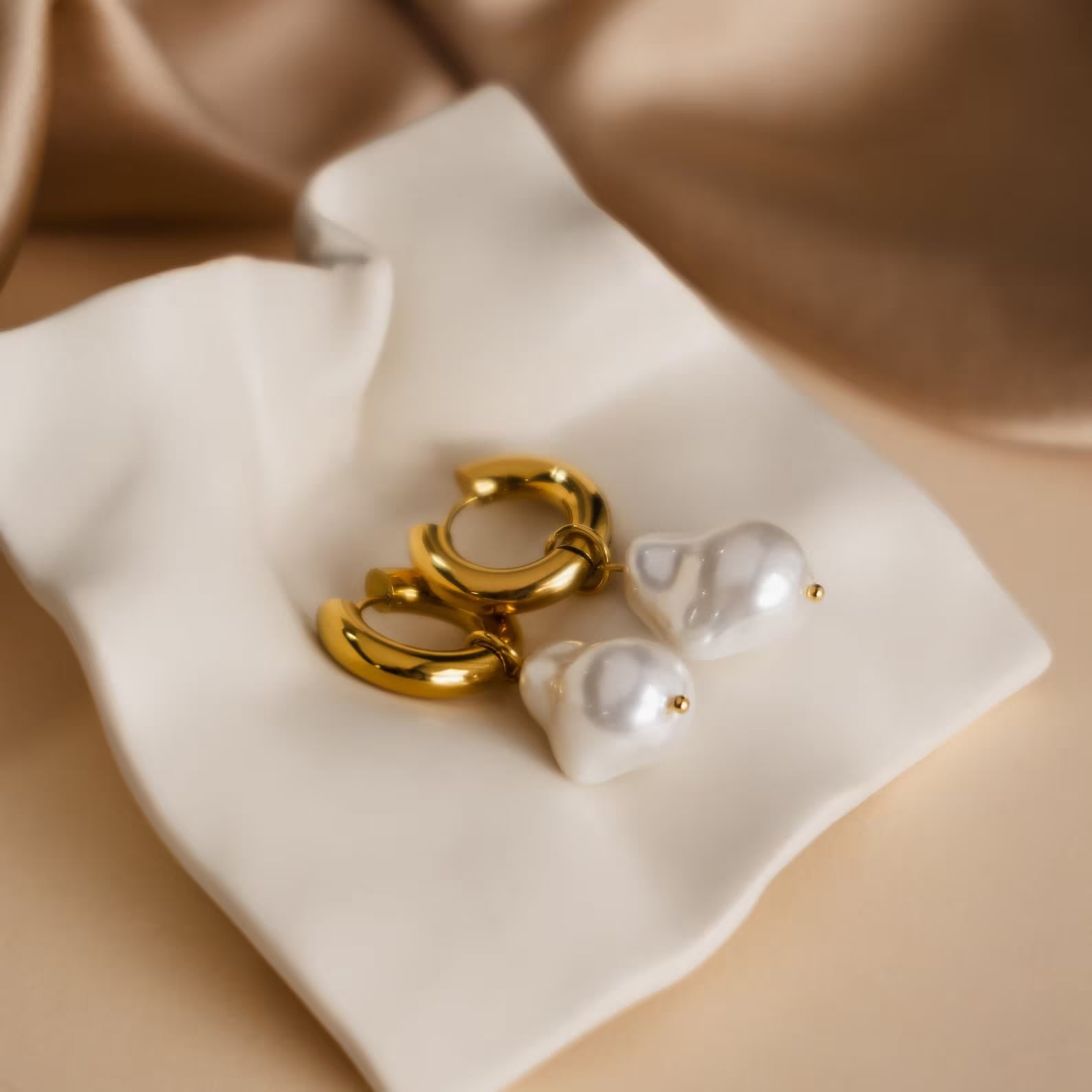 Baroque Pearl Earrings Set—gold hoops with dangling white pearls—rests elegantly on cream-colored fabric.