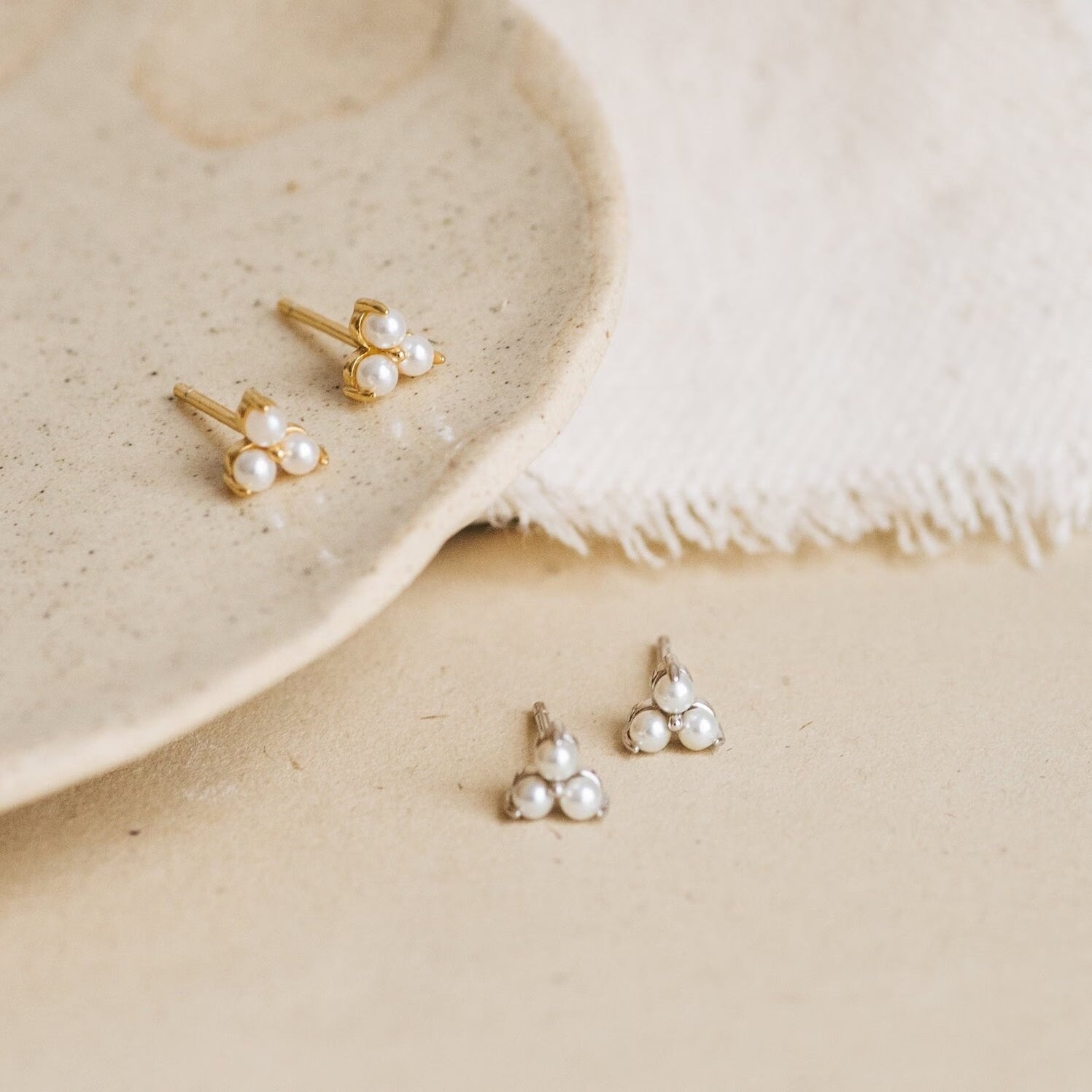The Baroque Pearl Earrings Set, featuring gold and silver pearl studs on a ceramic plate and fabric, is ideal for mixing with baroque pearl earrings or layering with delicate pearl drop hoops.