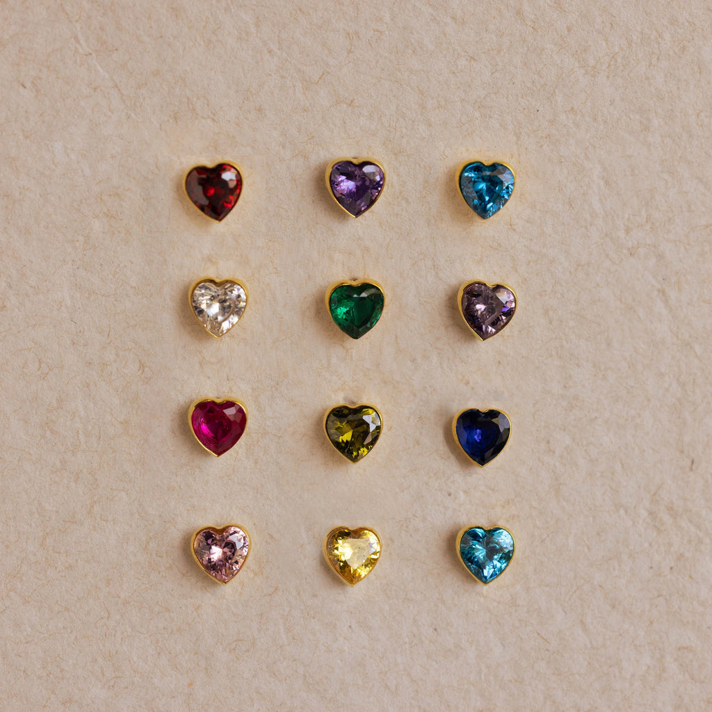 Twelve Bezel Heart Birthstone Studs, each featuring a multicolored gemstone, are arranged in a neat grid on a beige textured surface.