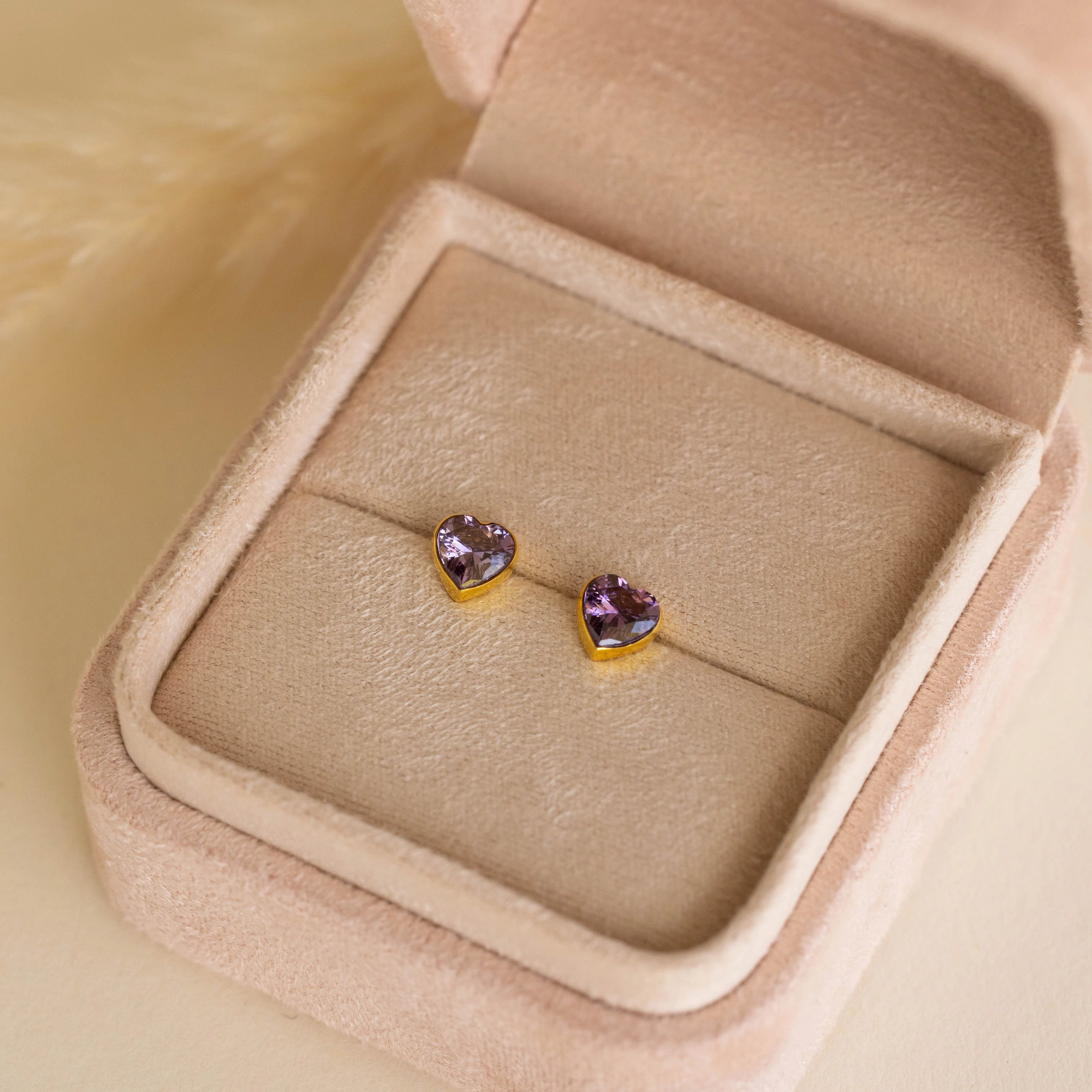 The Bezel Heart Birthstone Studs, featuring heart-shaped purple gemstones, are elegantly presented in a beige velvet jewelry box.
