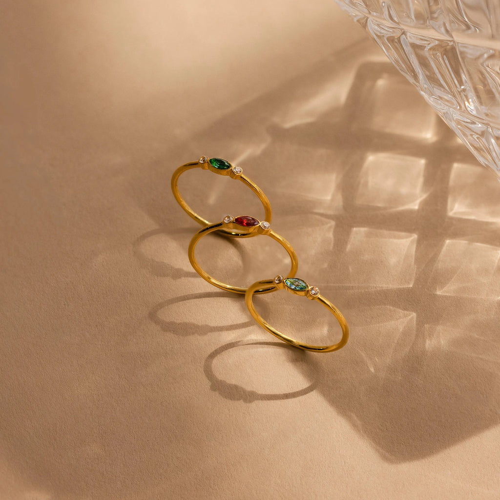 The Marquise Bezel Birthstone Ring, a customizable gold ring with colorful gemstones, rests on a beige surface beside a glass bowl casting a shadow.